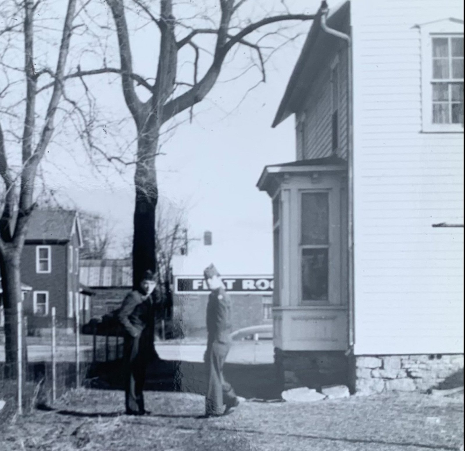 A black and white photo of children playing on a playground with a tree and houses in the background. A sign that partially reads 'FEST R' is visible.