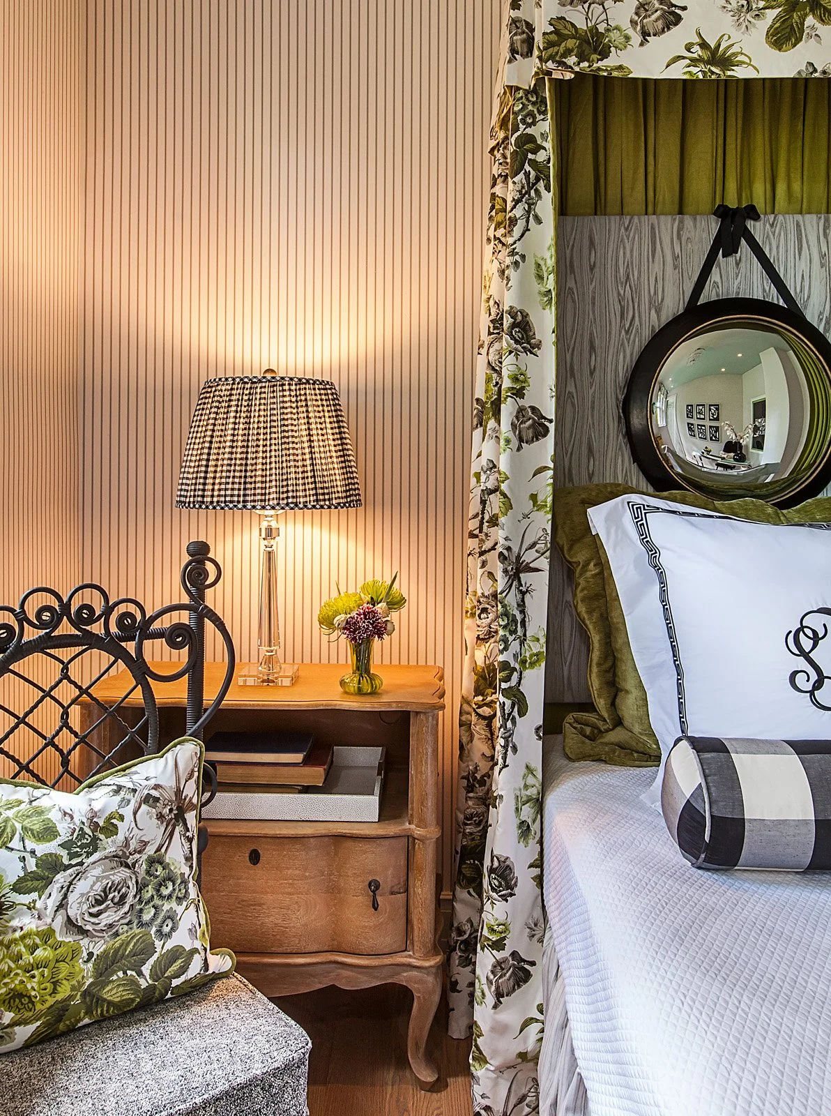 A cozy bedroom with a lamp on a wooden nightstand, a colorful flower arrangement, a floral patterned pillow, a bed with white and dark green bedding, an oval mirror hanging above the bed, and floral curtains.