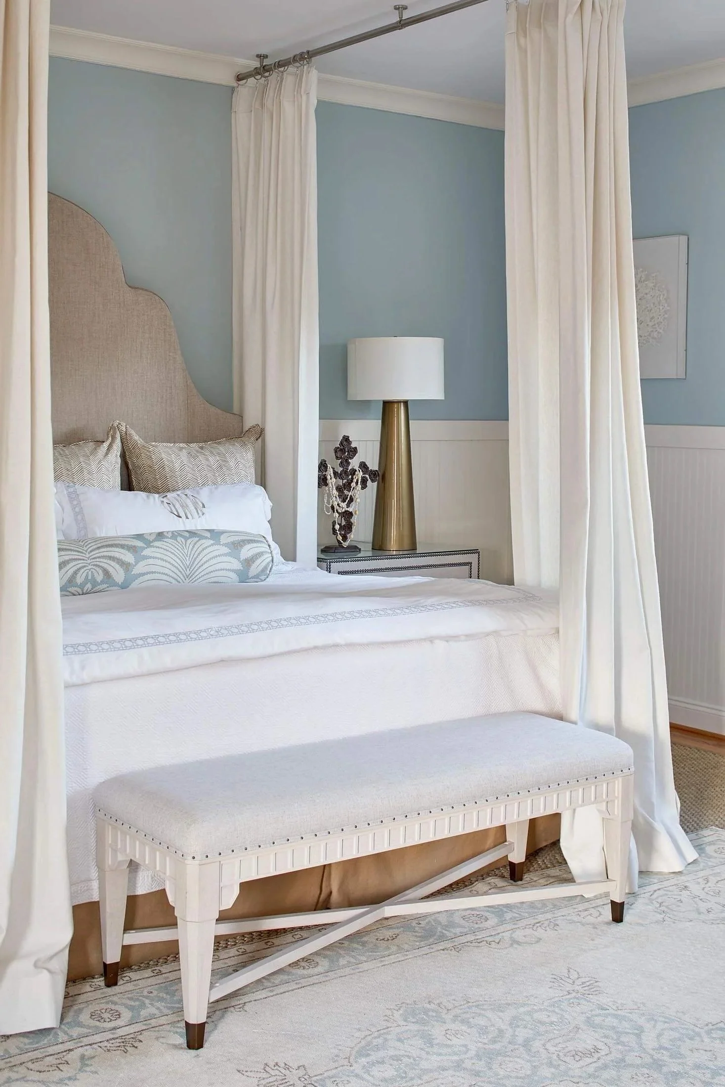 A bedroom with a bed featuring a beige headboard, white bedding with patterns, and decorative pillows. There are white curtains around the bed, a gold and white table lamp, a piece of artwork on the wall, and a white bench at the foot of the bed. The room has light blue walls and a patterned rug on the floor.