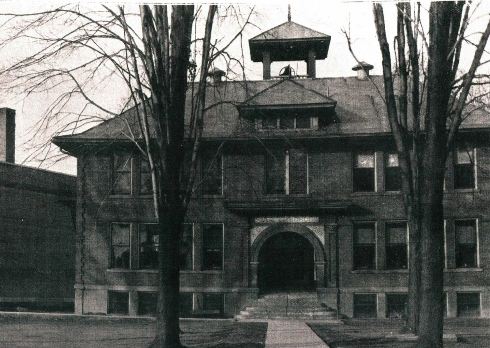 Black and white photo of a large, old brick building with arched entrance, multiple windows, and a prominent tower with a pyramid-shaped roof on top. Two leafless trees are in the foreground.