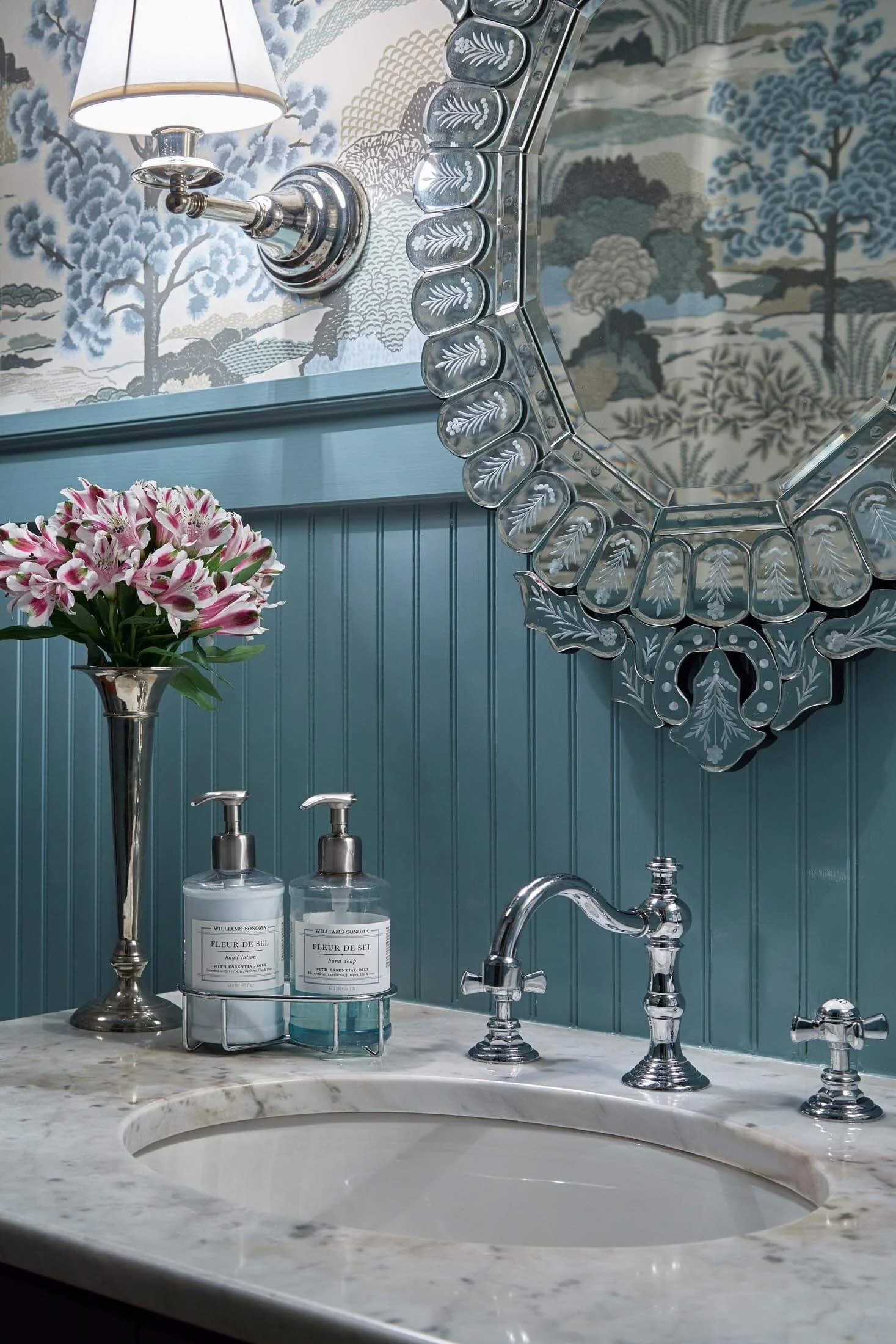 A bathroom vanity with a marble countertop, a silver faucet, and two bottles of hand soap labeled 'Fleur de Sel.' A floral arrangement of pink and white flowers in a silver vase is beside the soap. A decorative mirror with an octagonal shape and etched floral patterns hangs above a blue shiplap wall. A wall-mounted light fixture with a white shade is reflected in the mirror, along with wallpaper featuring a blue and gray landscape scene with trees and rolling hills.
