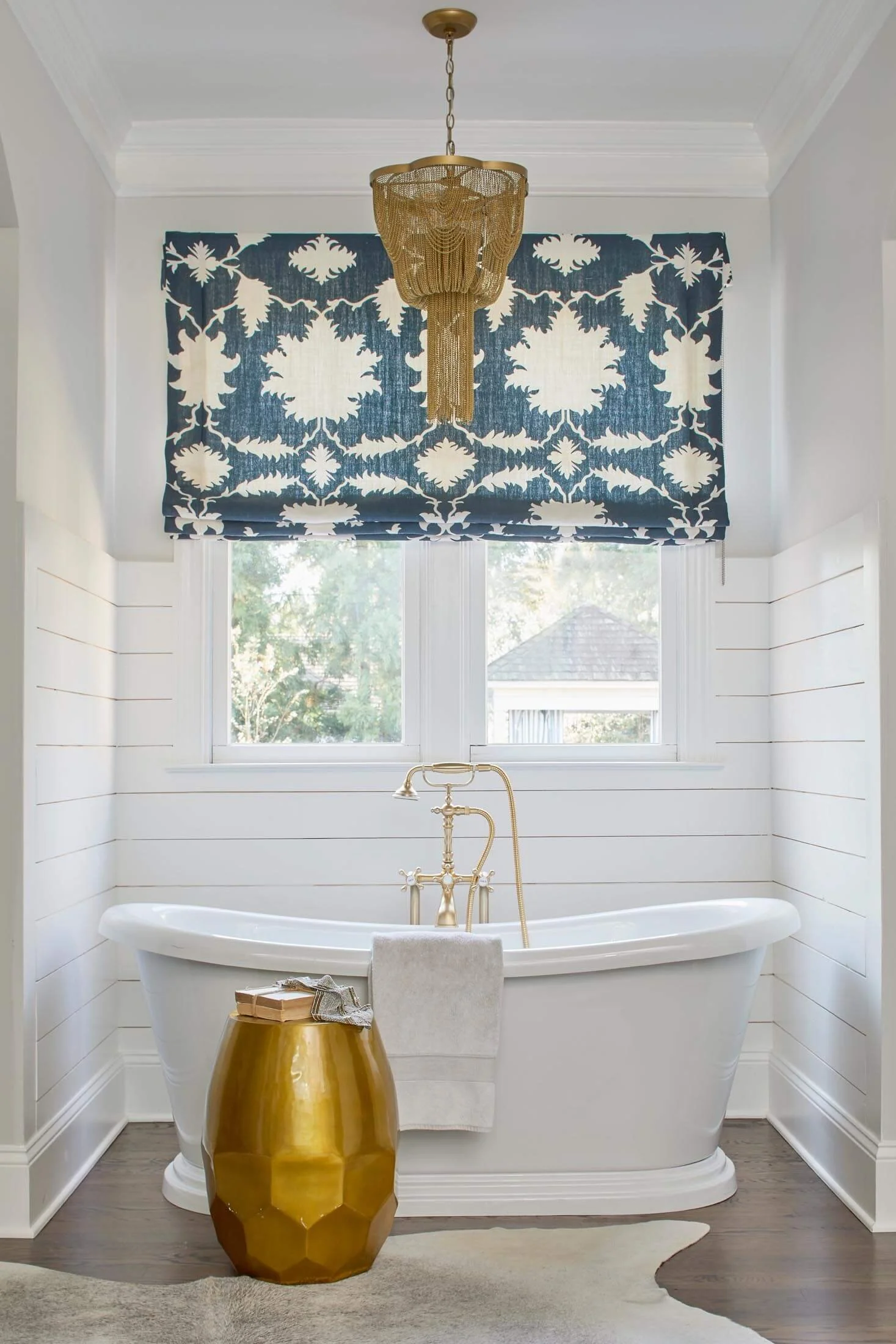 A modern bathroom with a white clawfoot bathtub, a gold side table, a decorative gold faucet, and a large window with a blue and white patterned Roman shade. A gold chandelier hangs from the ceiling.