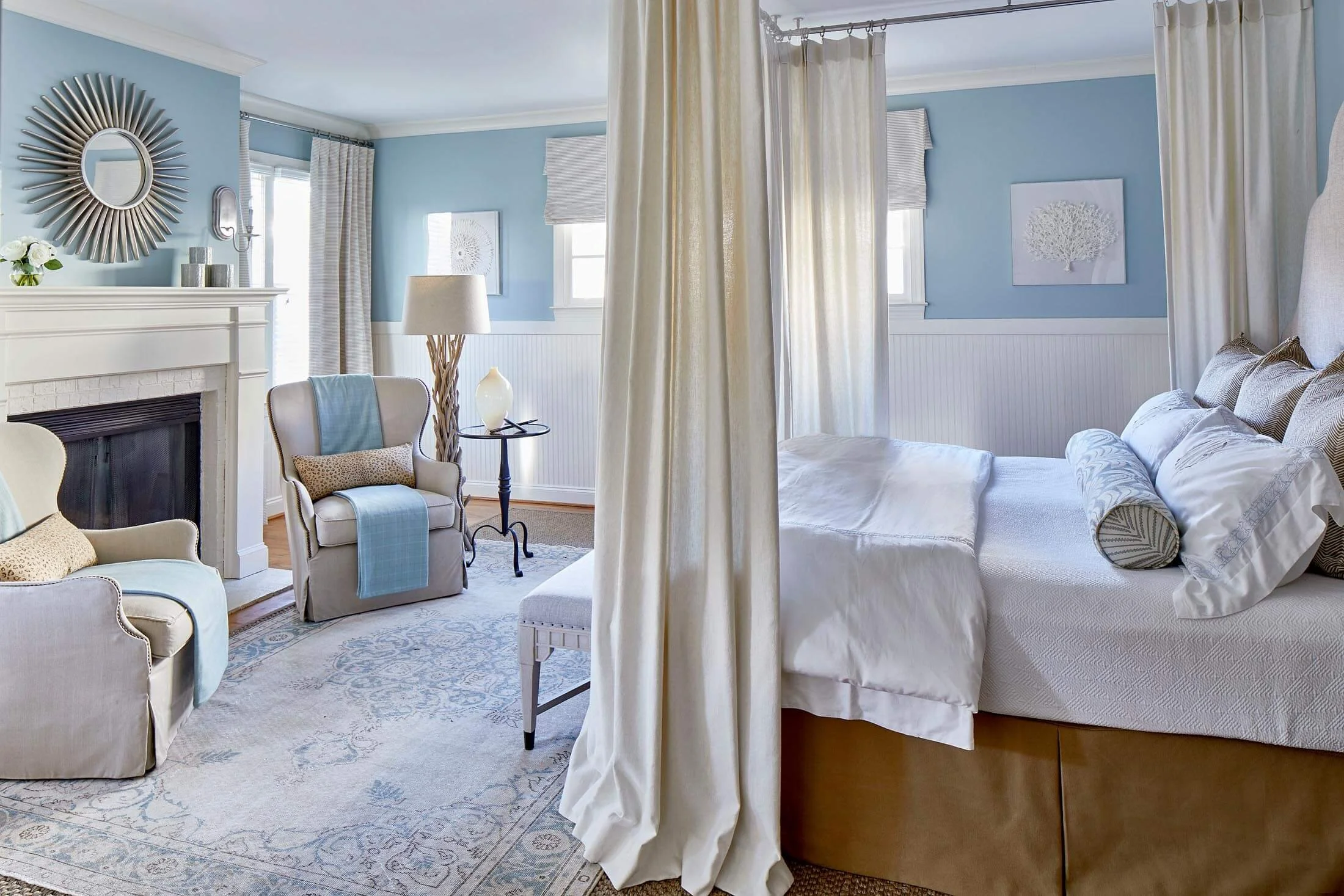 A cozy bedroom with a white bed, blue walls, and cream curtains, separated by a curtain rod. The room has vintage-style furniture, a fireplace with decorative items, and framed art wall decor.