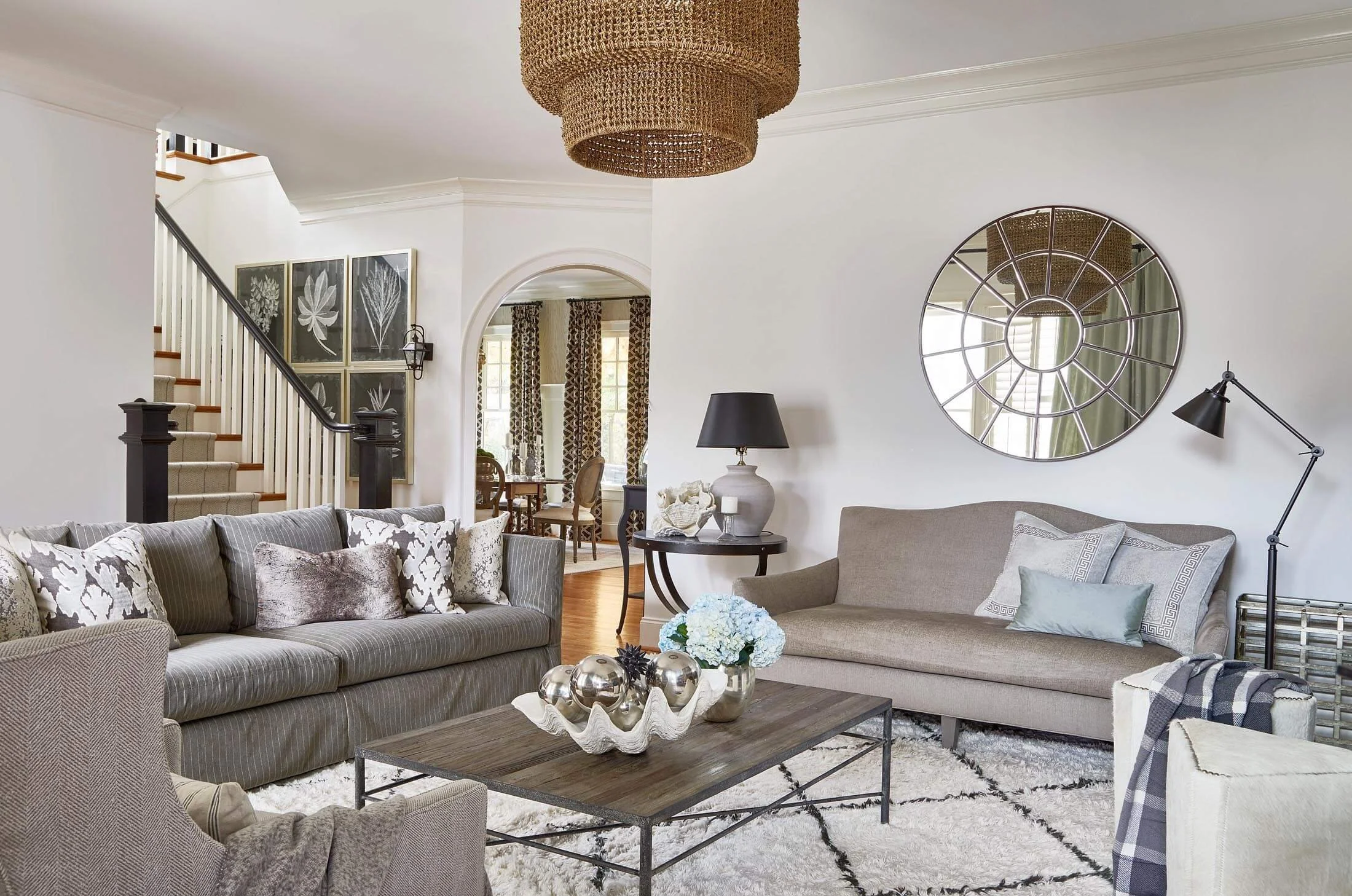 A living room with gray sofas, patterned pillows, a coffee table with decorative balls and flowers, a black lamp, a large mirror, wall art, and a staircase leading to an upper floor.