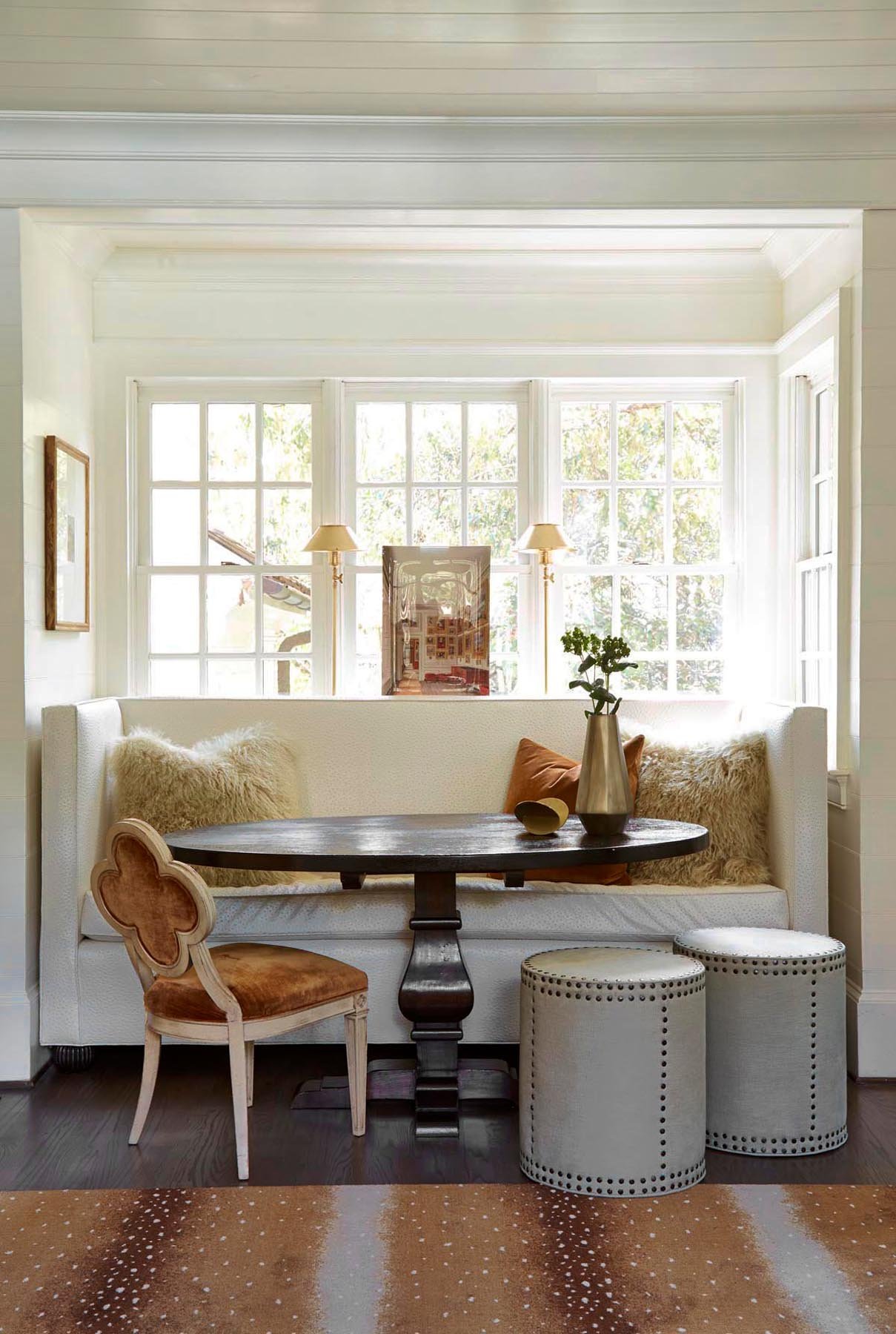 Bright living room with large window, white built-in bench with beige and brown pillows, a small wooden table, decorative vases, and furniture including a pink upholstered chair and white textured stools. Rug with brown and white pattern on dark wood floor.