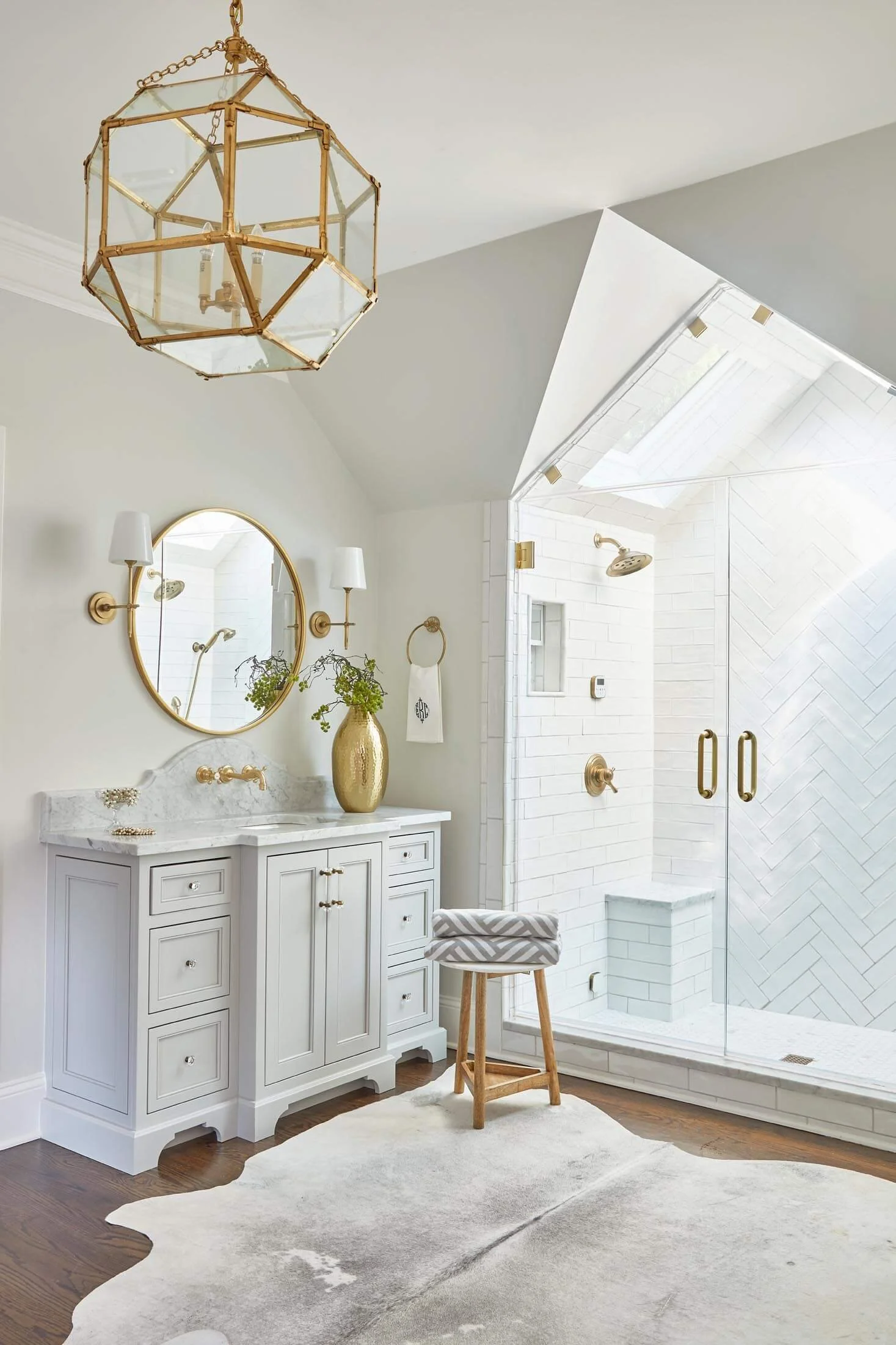 Bright bathroom with white cabinetry, a gold-framed mirror, brass sconces, a gold vase with greenery, a modern stool, a large cowhide rug, and a glass-enclosed shower with white subway tile and a skylight.
