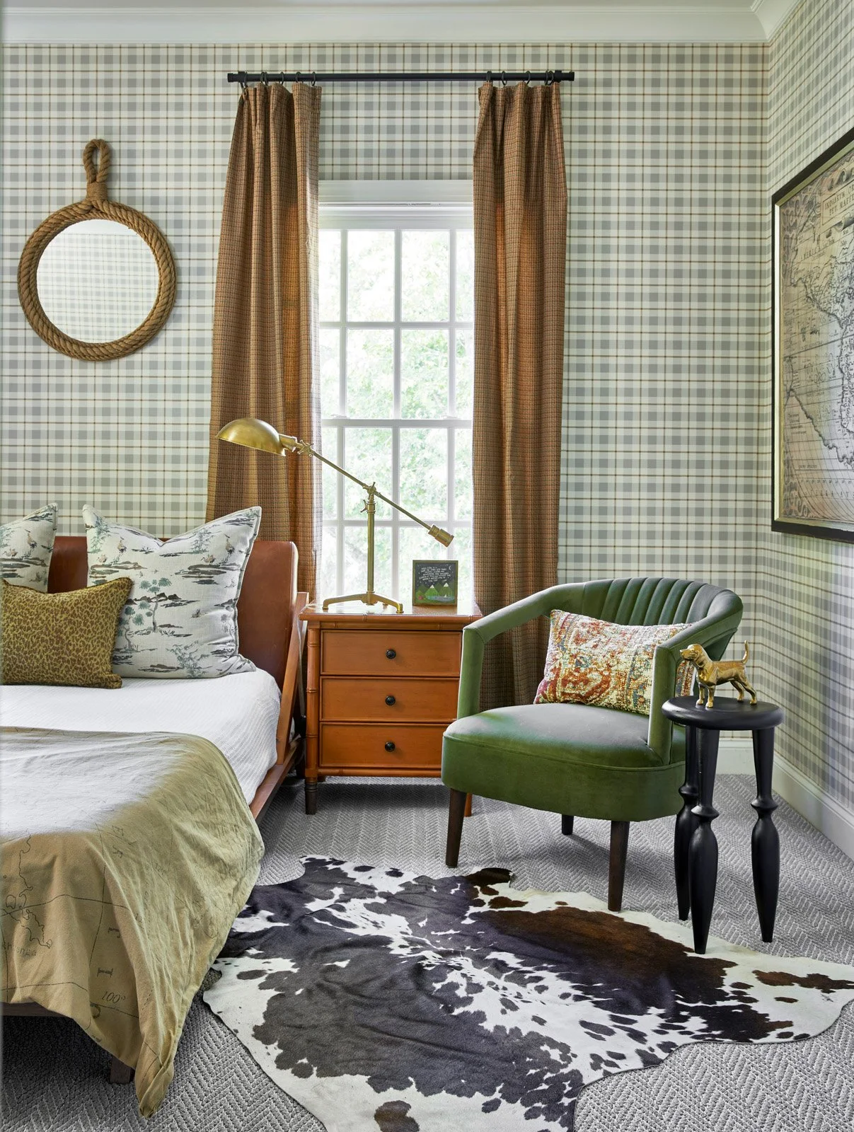 A cozy bedroom corner with plaid wallpaper, beige curtains, a wooden bed with patterned pillows, a wooden nightstand with a brass desk lamp, a green upholstered armchair with a decorative pillow, a small black side table with a gold dog figurine, and a cowhide rug on a grey carpeted floor.