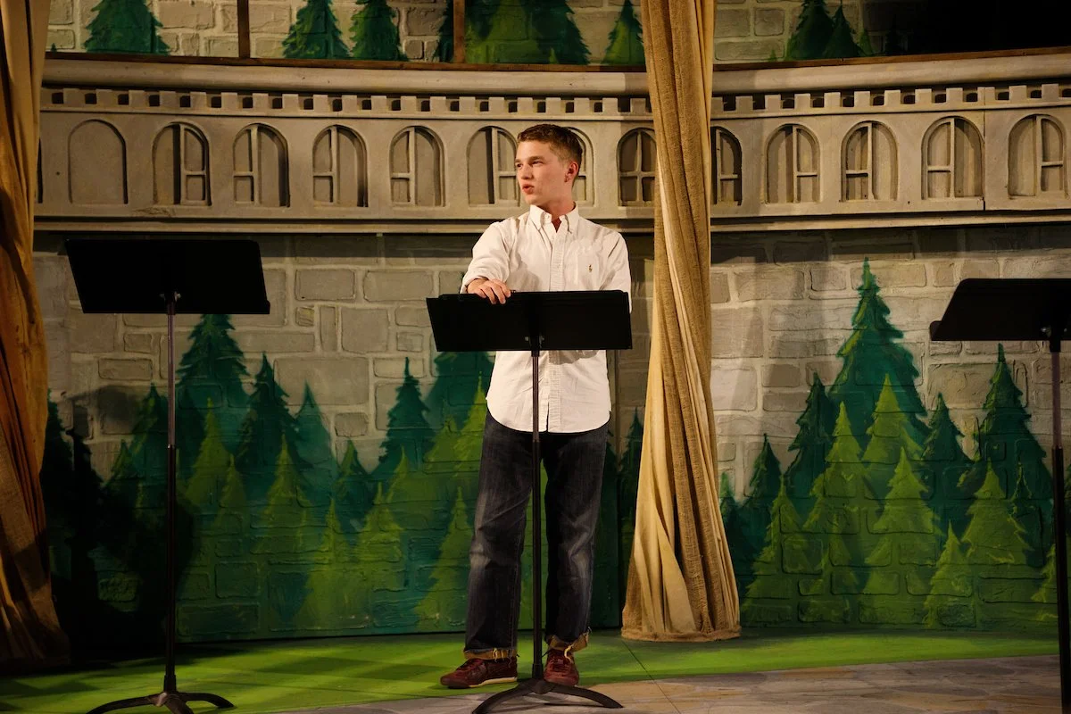 A young man stands on stage behind a music stand, with a painted forest background and gold curtains on either side.