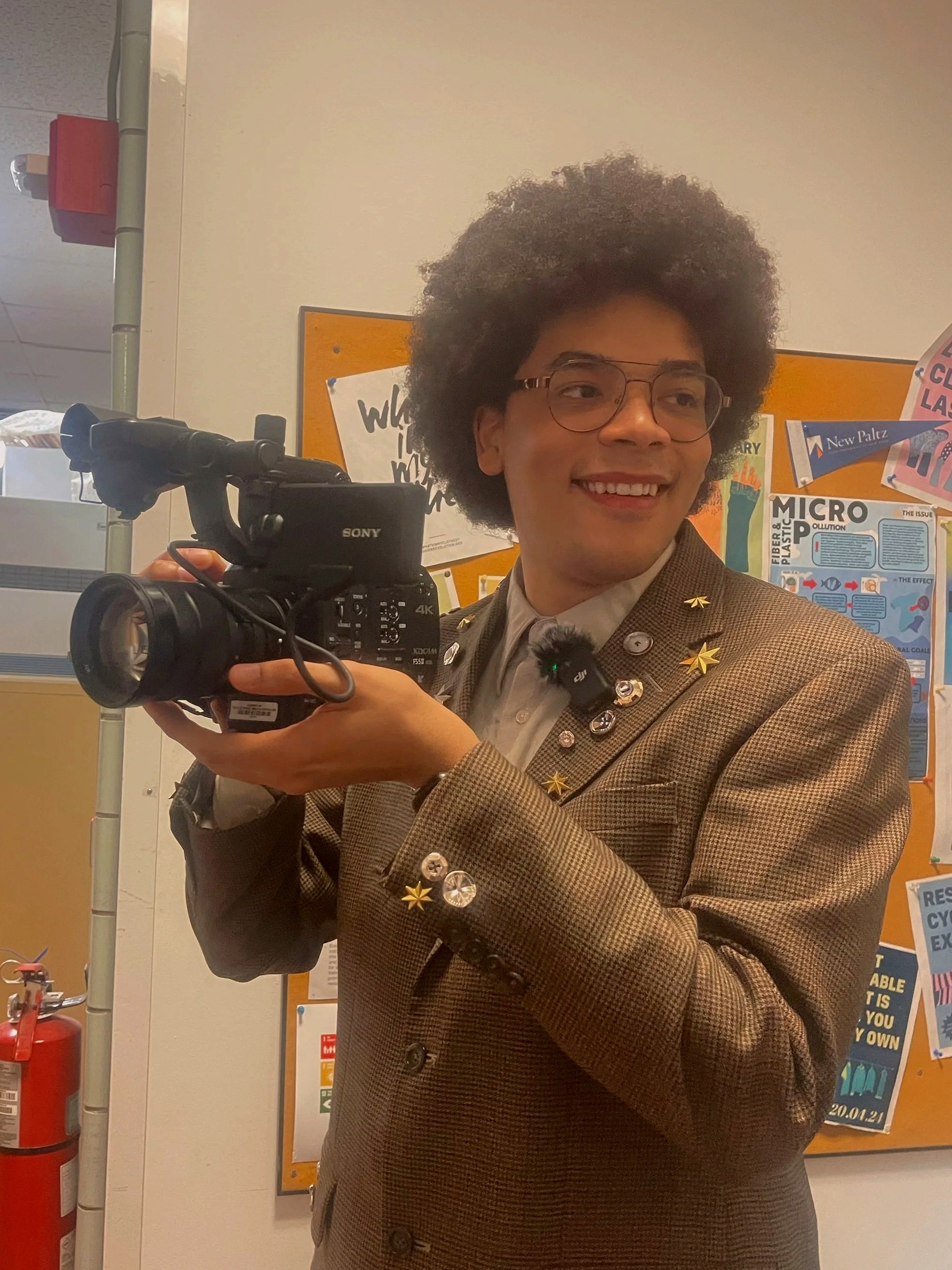 A person with glasses and an Afro hairstyle, smiling and holding a camera, dressed in a brown checkered blazer with star-shaped pins, standing in front of a bulletin board with posters about recycling and community events.