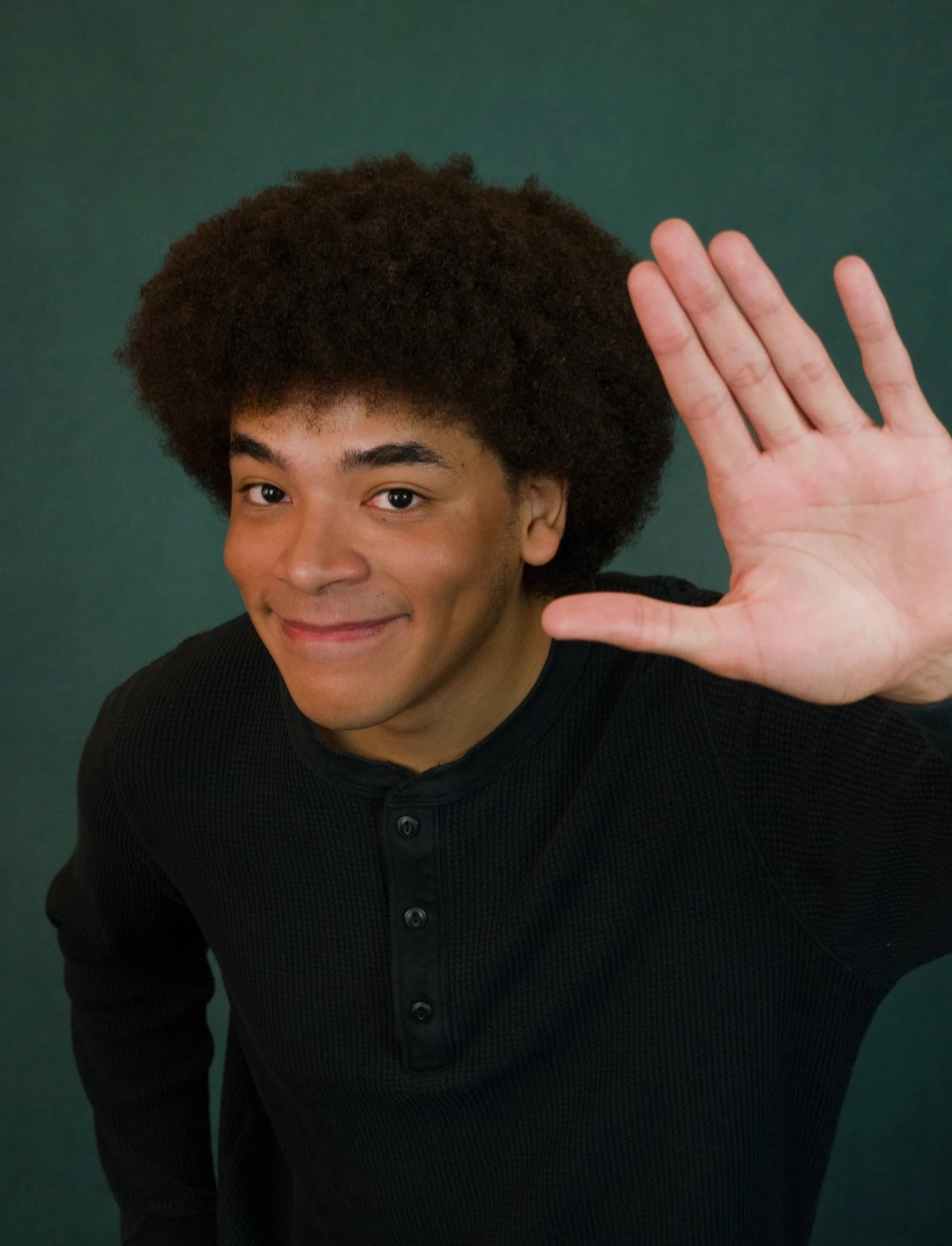 A young man with curly hair smiling and holding up his right hand in a greeting gesture against a green background.