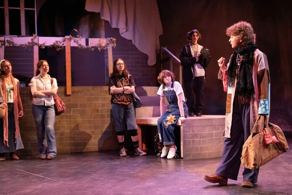 A group of young women watching a woman speak on stage in a theatrical production.