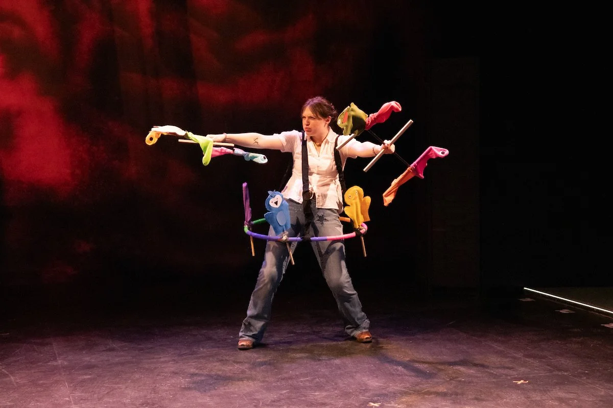 Performer on stage wearing a costume with plush toy arms attached to a harness, holding sticks, with a dark background and red stage lighting.
