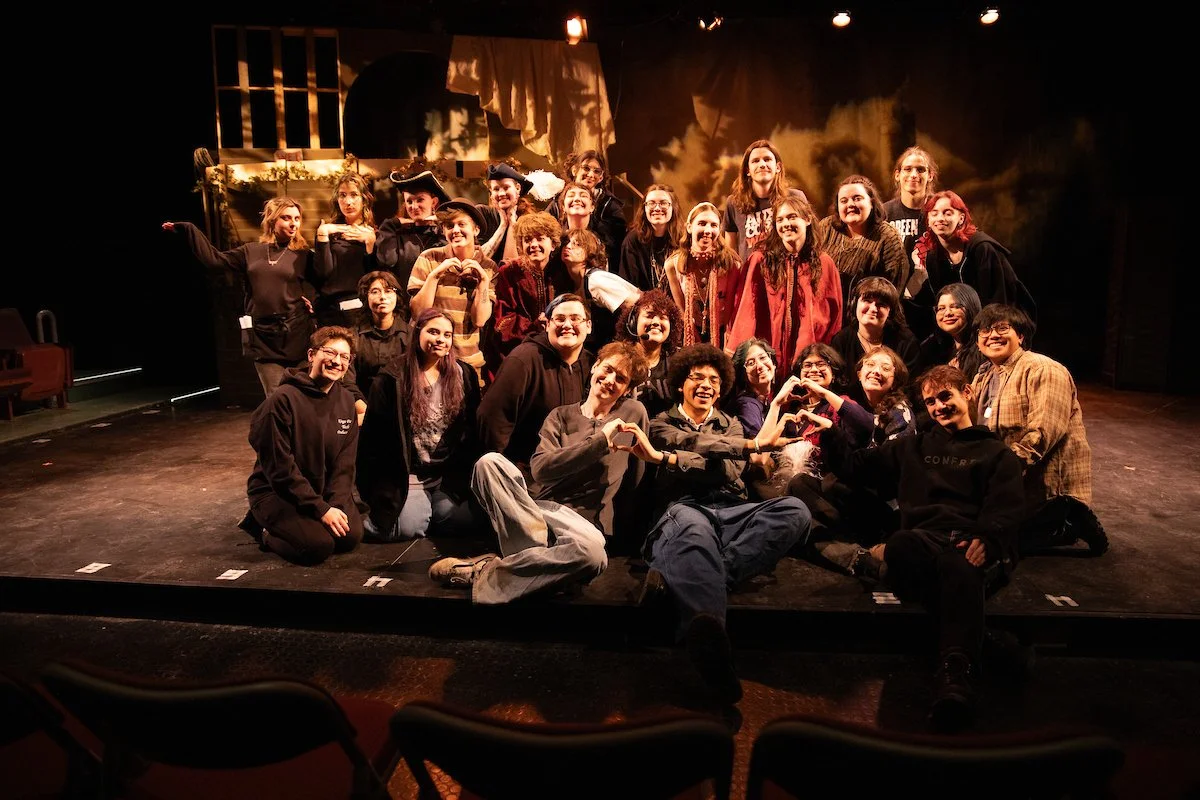 Group of young adults and teens on stage, some dressed in costumes, posing for a photo after a theater performance. They are smiling, making heart shapes with their hands, and standing in front of a theater set with dark lighting.