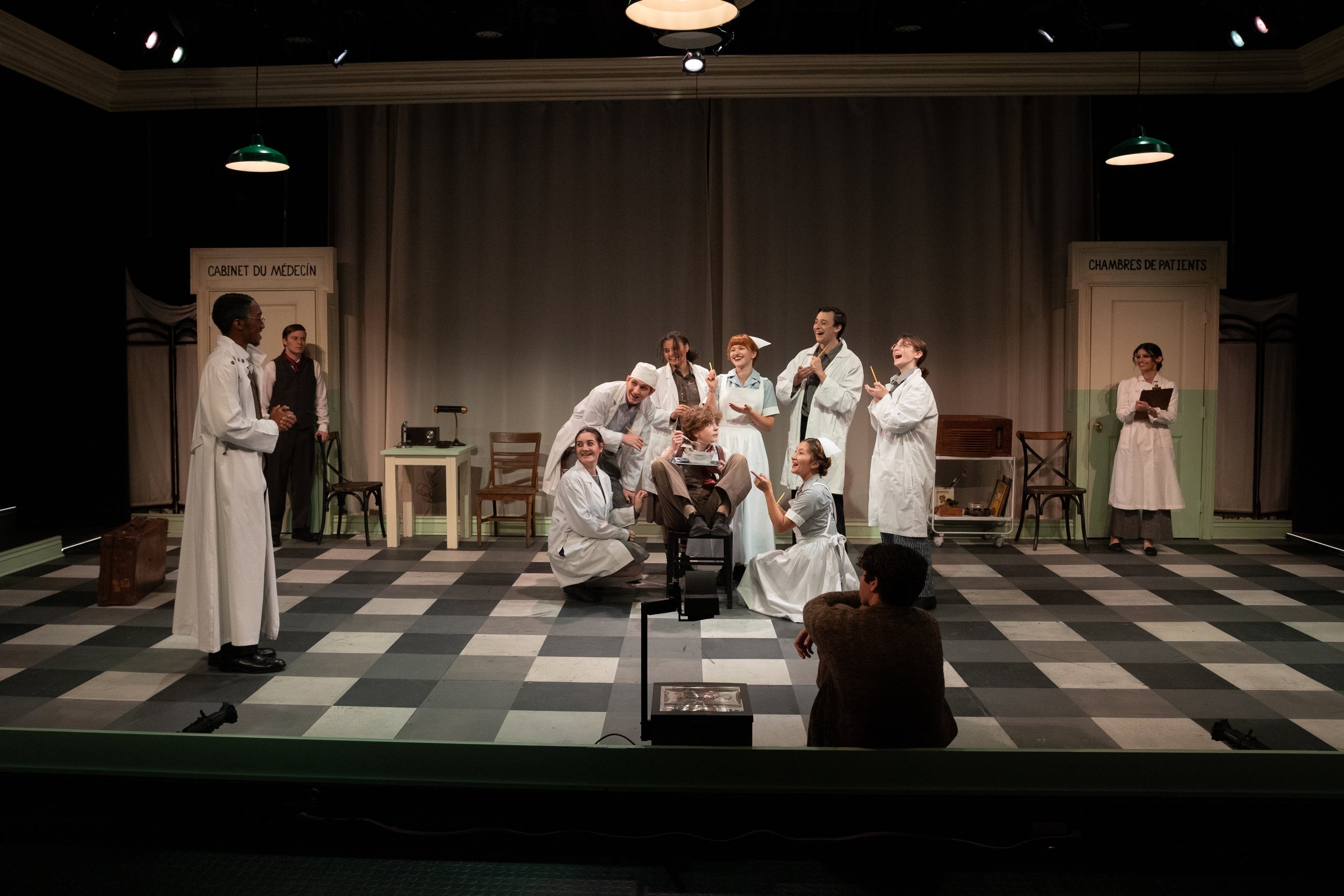 Theater stage set as a hospital ward with actors in medical costumes performing a play; villagers are on stage, some sitting and some standing, with medical signs background.
