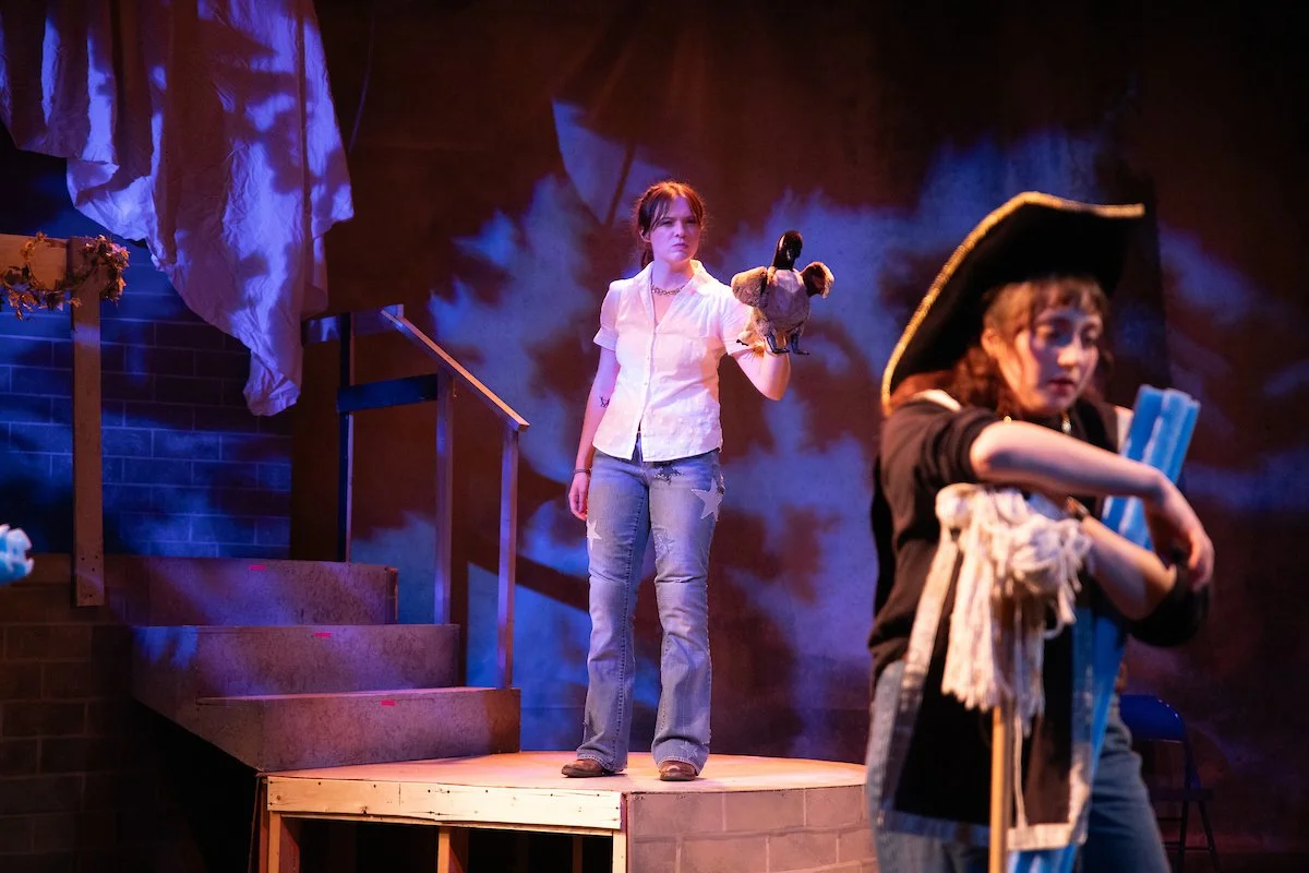 Two women performing on a stage with theatrical lighting and scenery, one holding a stuffed animal and the other wearing a pirate hat.