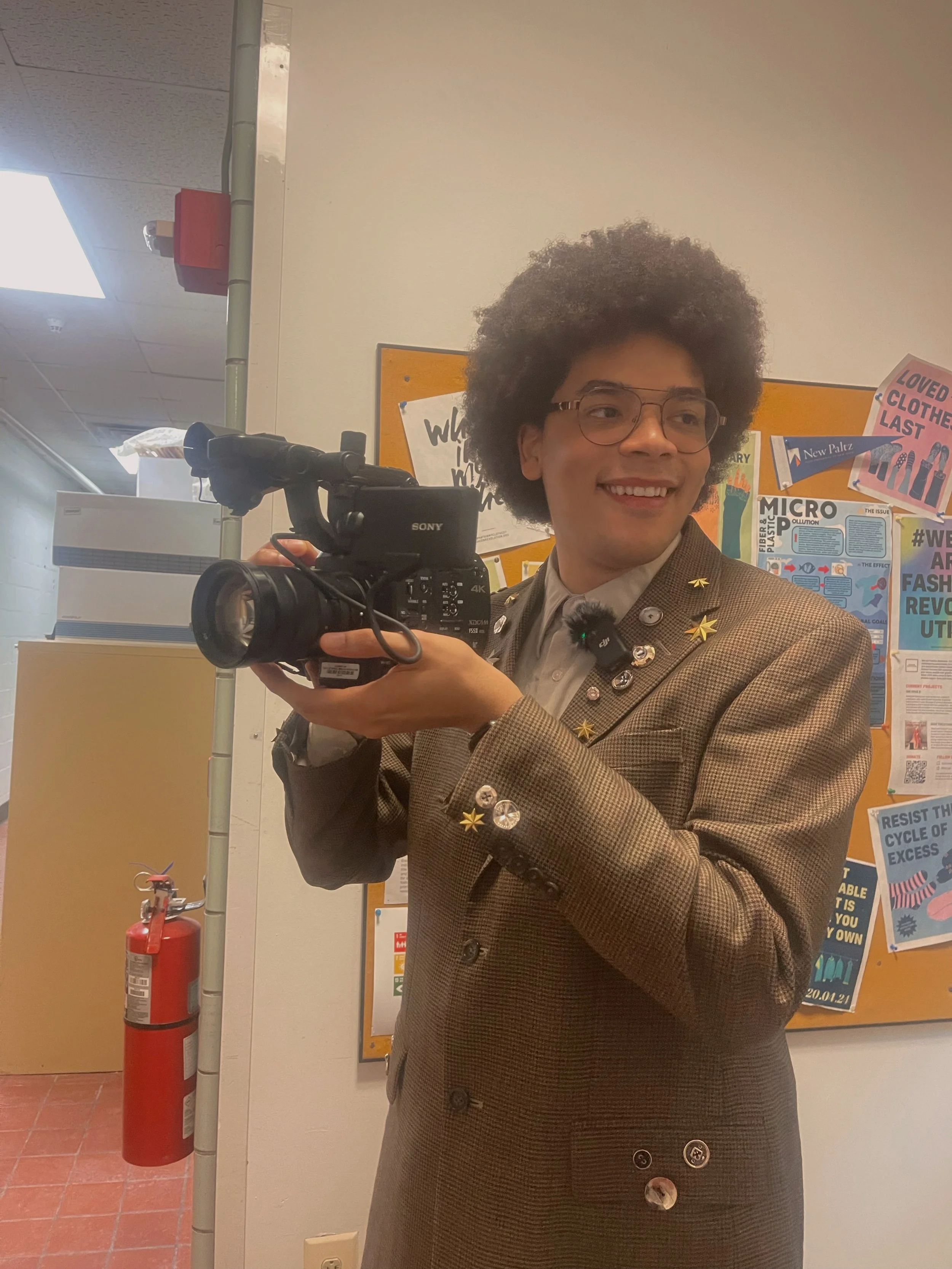 Person holding a video camera, wearing a brown blazer with pins, standing indoors with a bulletin board in the background.
