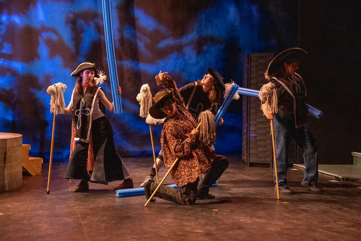 Four actors on stage dressed as pirates, holding blue plastic tubes, with one actor kneeling in front, set against a dark, cloudy backdrop.