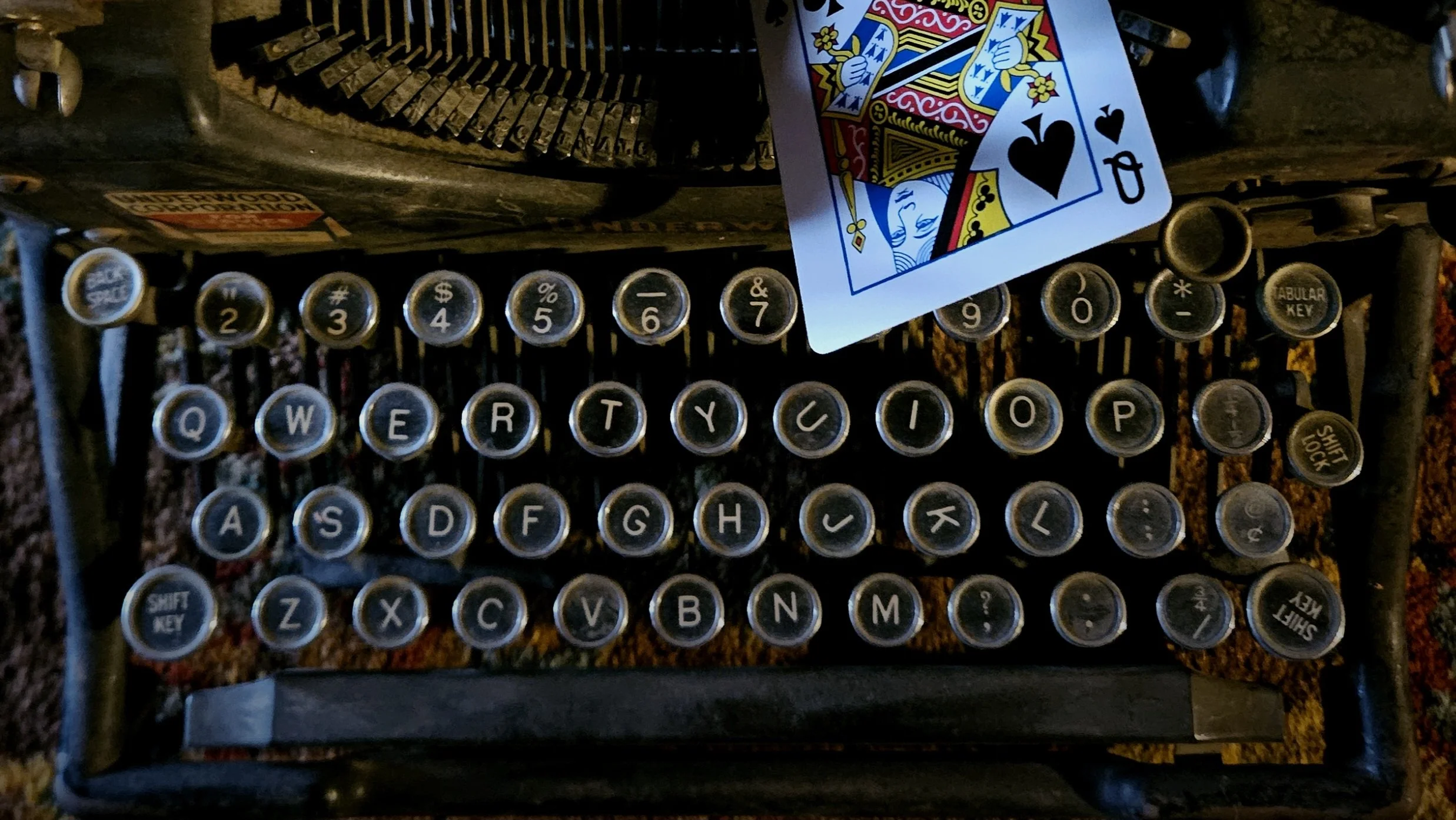 Close-up of a vintage typewriter raked in shadow, with a playing card (queen of spades) resting on it.