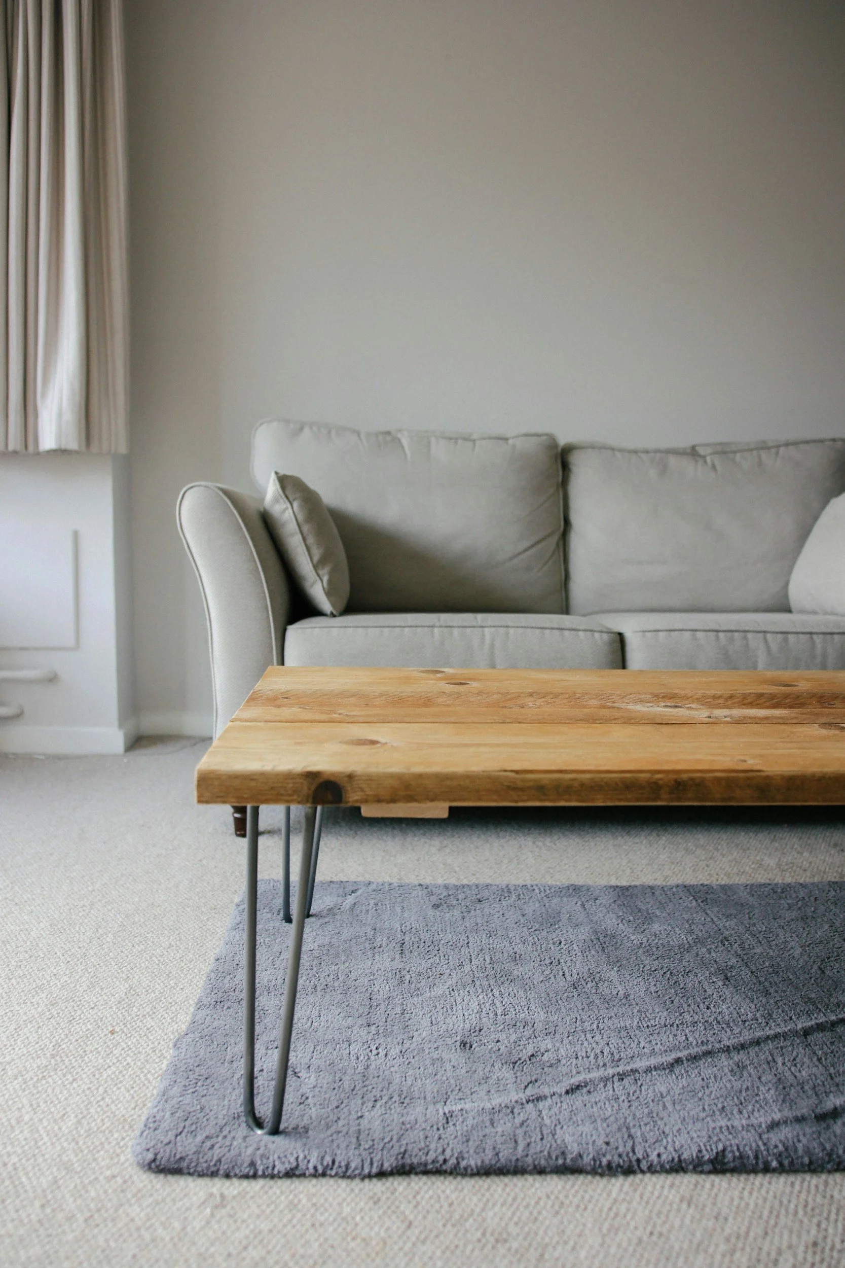 Living room with a gray sofa, a wooden coffee table, a small gray rug, beige carpet, and light-colored curtains.