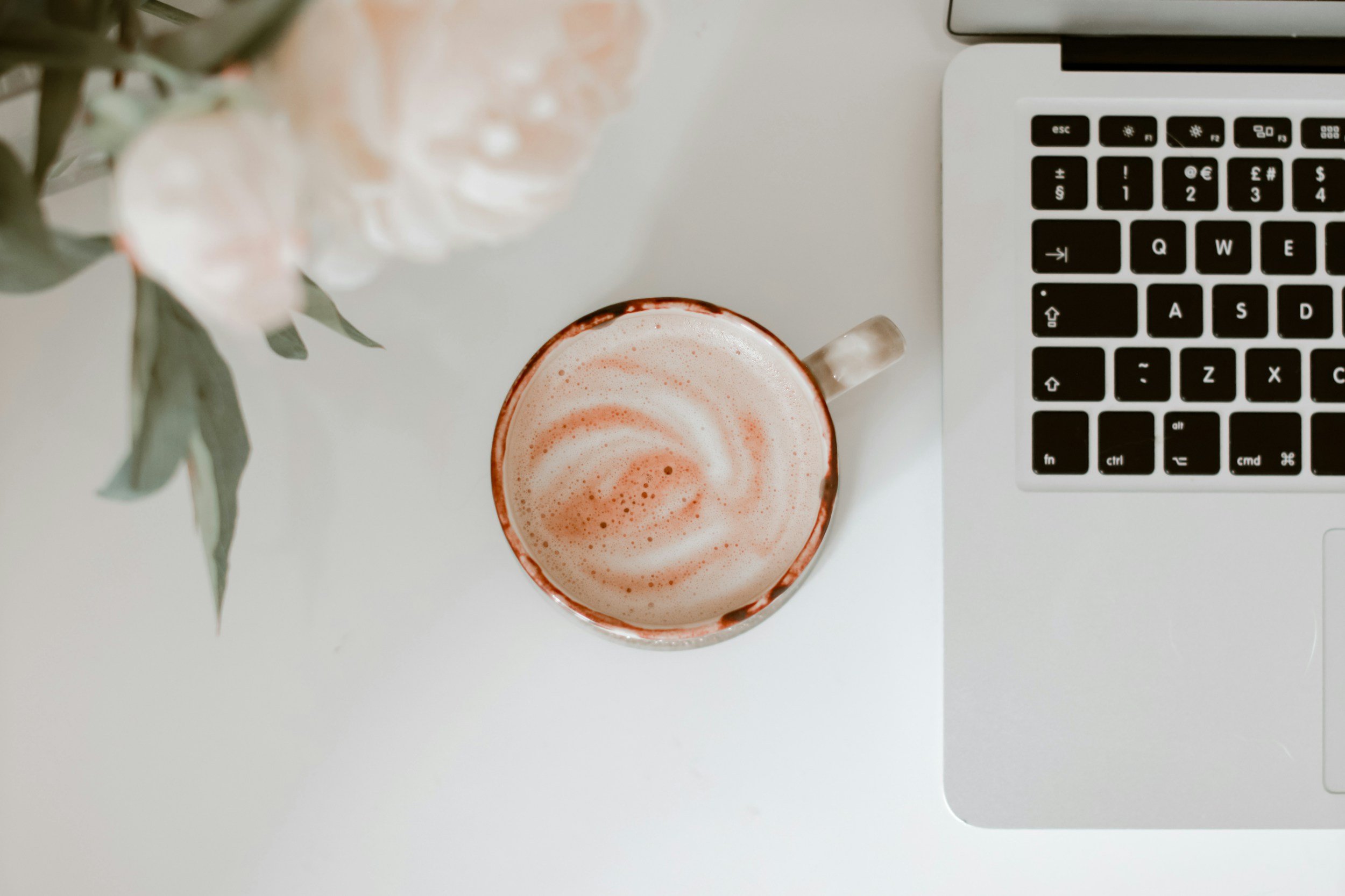Top view of a computer workspace with a laptop, a coffee cup with frothy beverage, and a blurred flower arrangement.