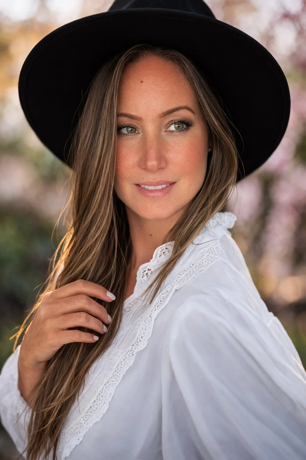 A woman with long light brown hair wearing a black hat and a white blouse with lace details, outdoors with blurred trees in the background.