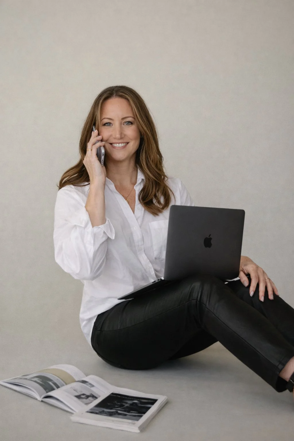 Woman sitting on the floor with a laptop on her lap, holding a phone to her ear, smiling, with an open magazine on the floor beside her.