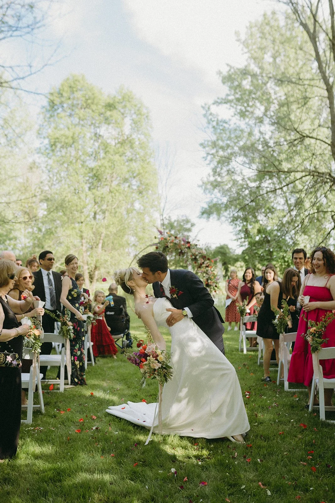 A bride and groom kiss during their outdoor wedding ceremony, surrounded by guests in a lush green park with trees. Rochester, new york wedding planning.