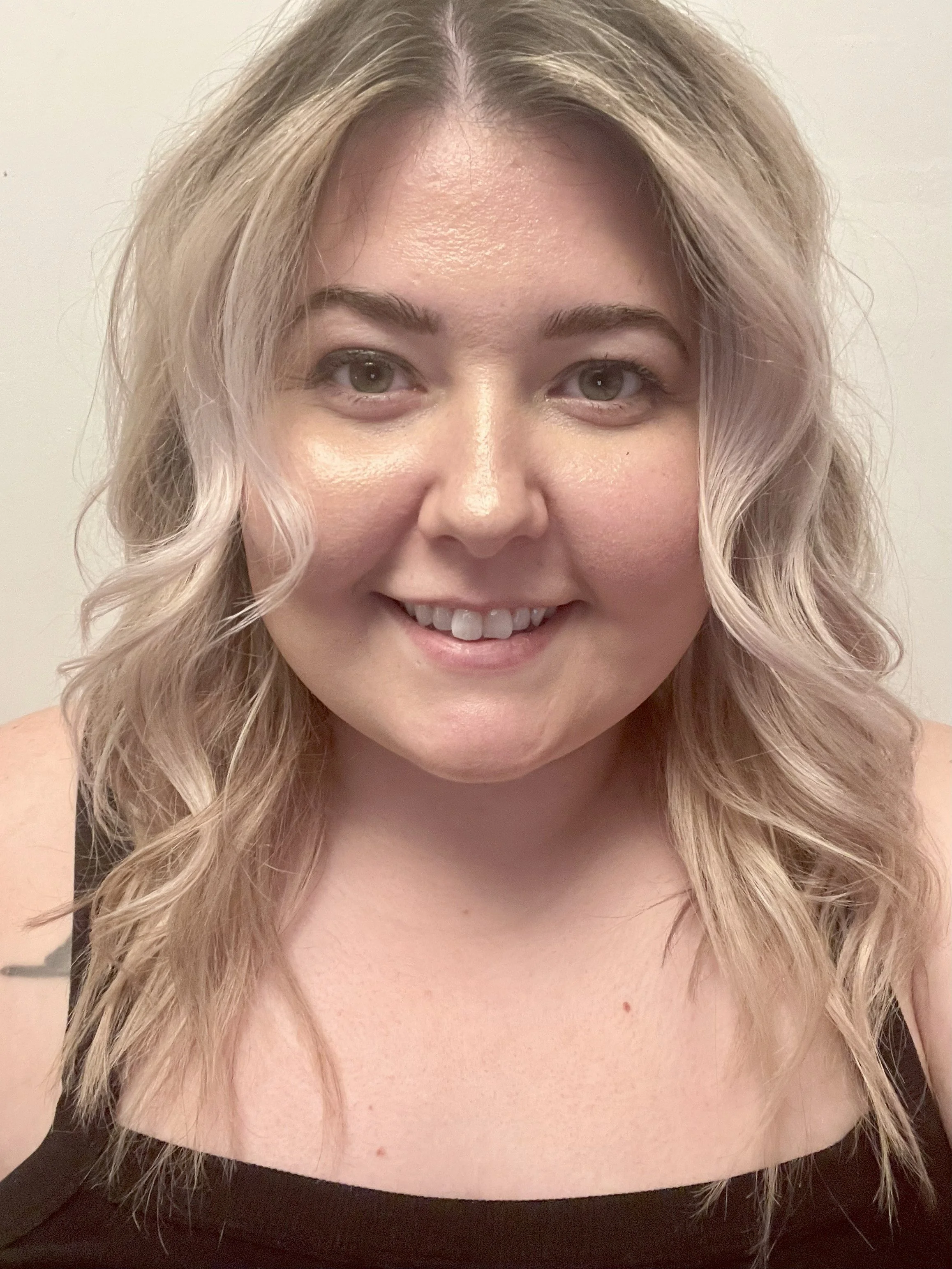 A woman with blonde, wavy hair smiling at the camera in front of a plain background.