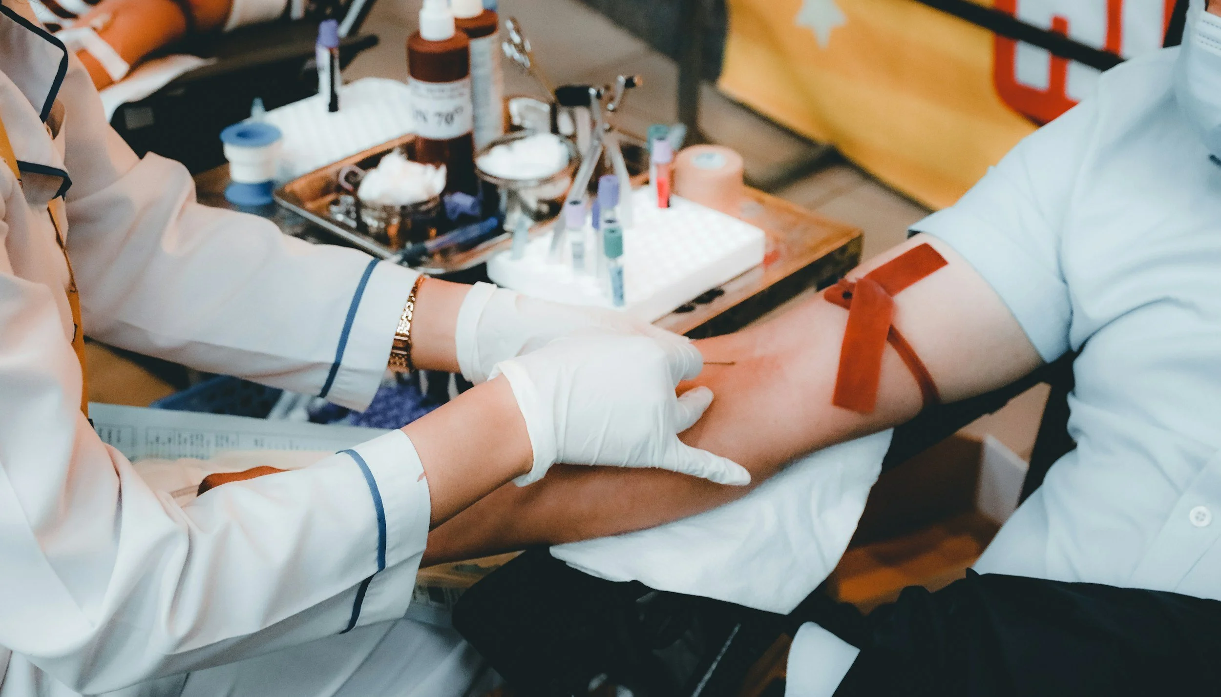 A person is donating blood while wearing a white shirt. Medical personnel are preparing to draw blood, with medical equipment and supplies visible on a table in the background.