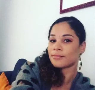 A woman with dark hair in a side ponytail, wearing earrings and a gray sweater with black spots, sitting on a couch. There is a white wall and a framed picture in the background.