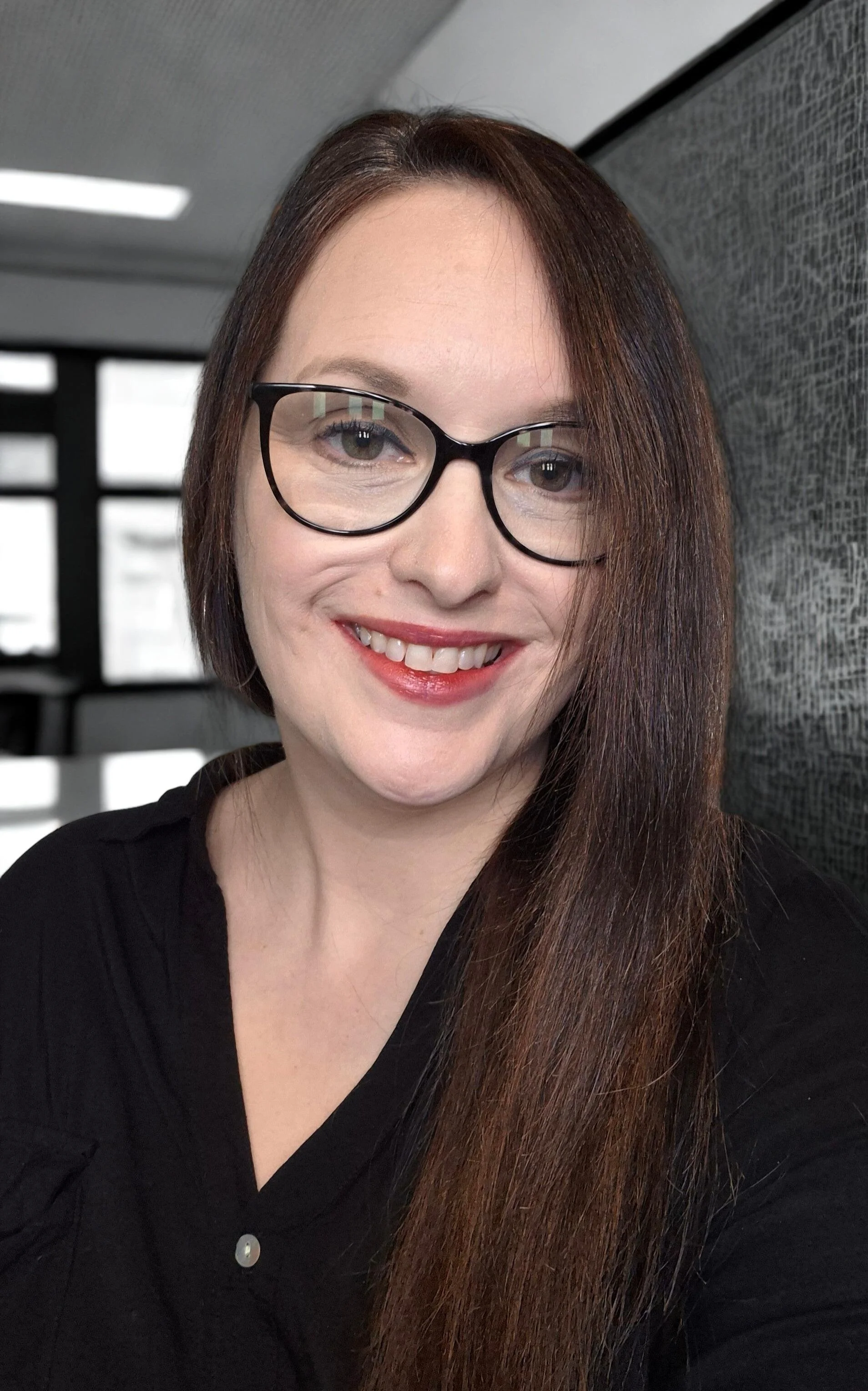 A woman with long reddish-brown hair, glasses, and a black top, smiling in an indoor setting with patterned wall and windows in the background.