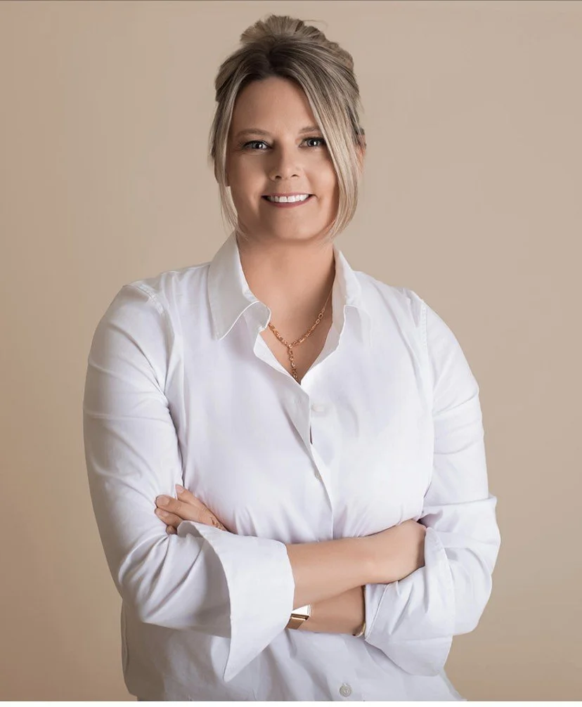 A woman with blonde hair styled in an updo, wearing a white button-up shirt and a gold chain necklace, standing with her arms crossed and smiling against a beige background.