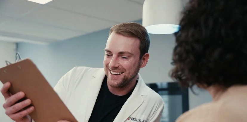 A man in a white coat smiling and holding a clipboard, engaging with a woman.