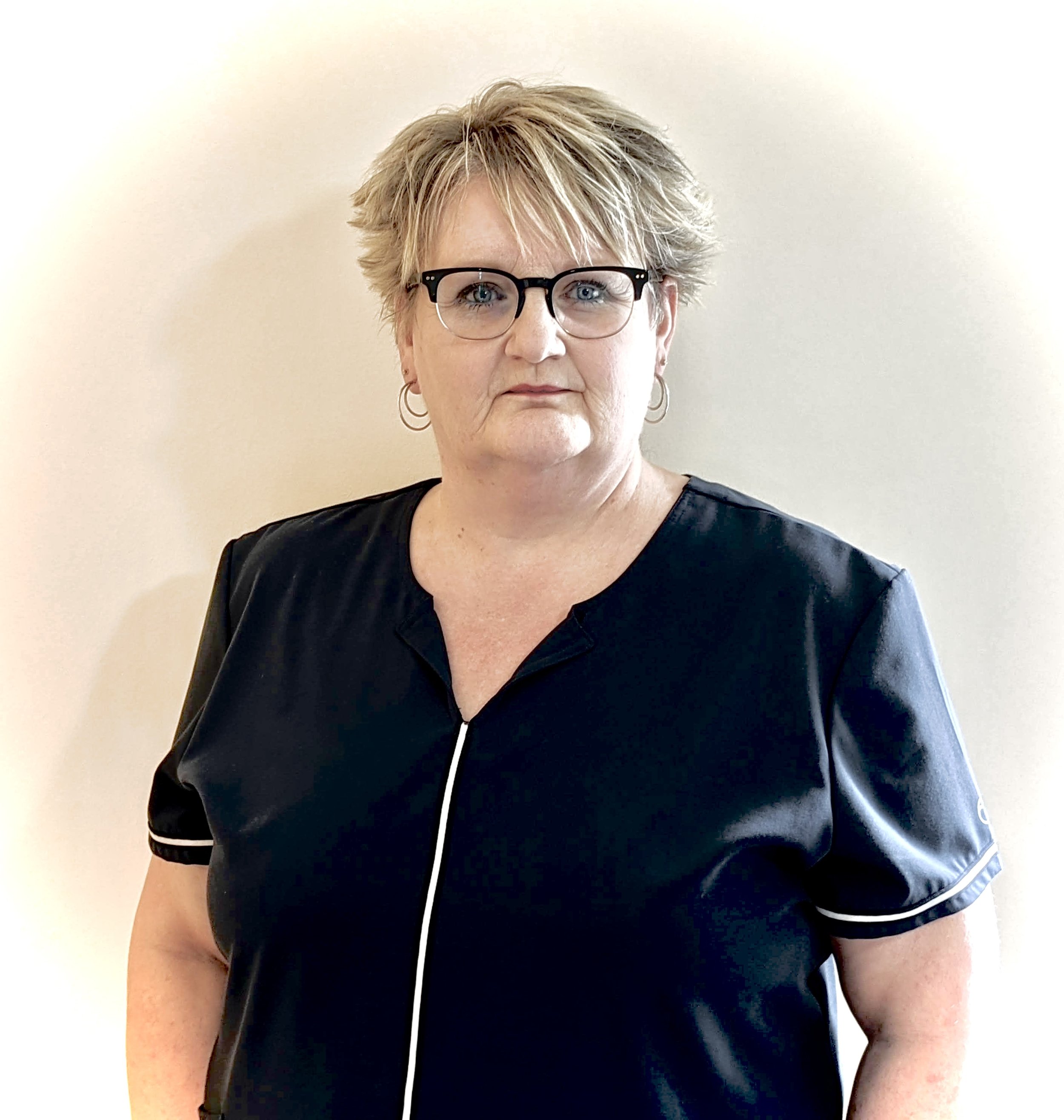 A middle-aged woman with short, tousled blonde hair, wearing black-rimmed glasses, hoop earrings, and a black top with white piping. She is standing against a plain light-colored wall, looking directly at the camera with a serious expression.