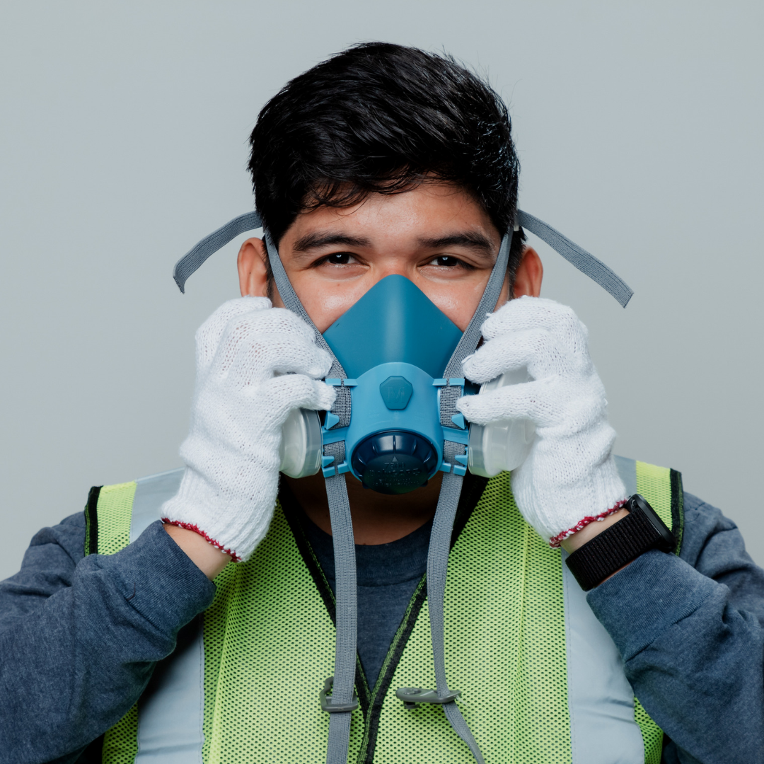 A man wearing a yellow safety vest, white gloves, and a black watch is holding a blue respirator mask in front of his face.