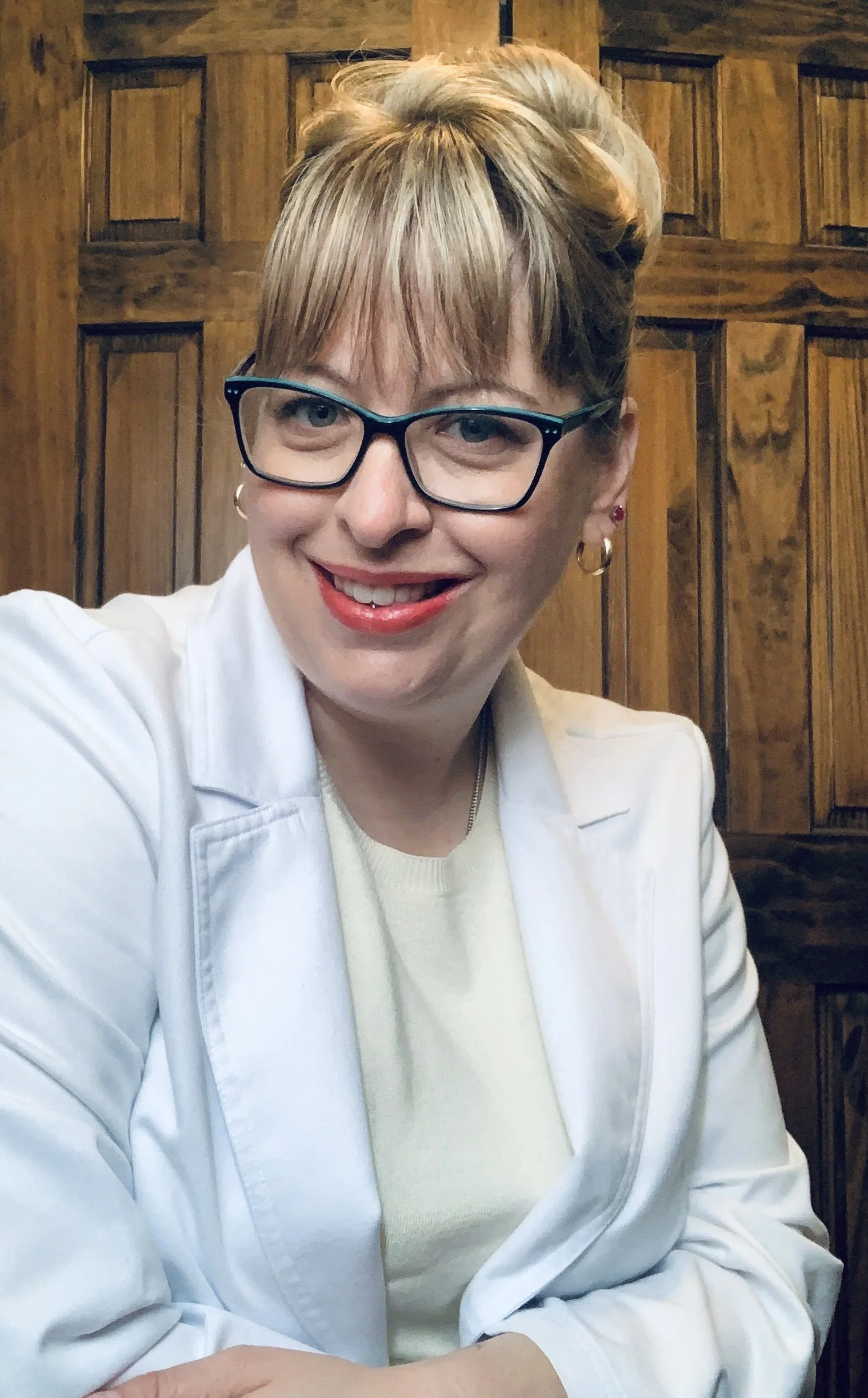 A woman with short blonde hair wearing glasses, a white blazer, and a light yellow top, smiling at the camera in front of a wooden background.