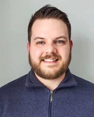 Headshot of a smiling man with dark hair and beard, wearing a navy blue quarter-zip sweater, against a light gray background.