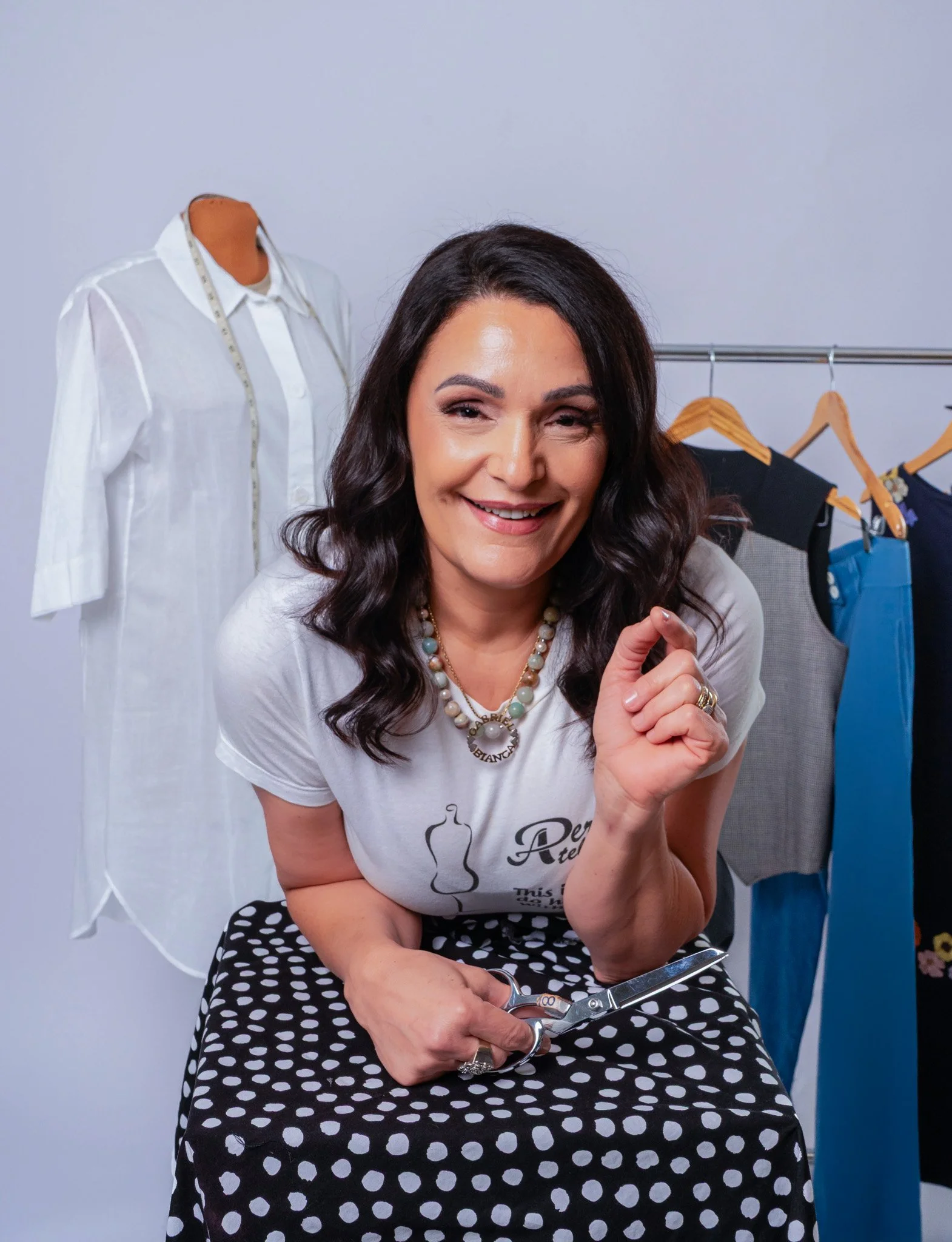 A woman smiling and holding scissors, sitting in front of a clothing rack with various garments including a white shirt, black pants, and blue pants.