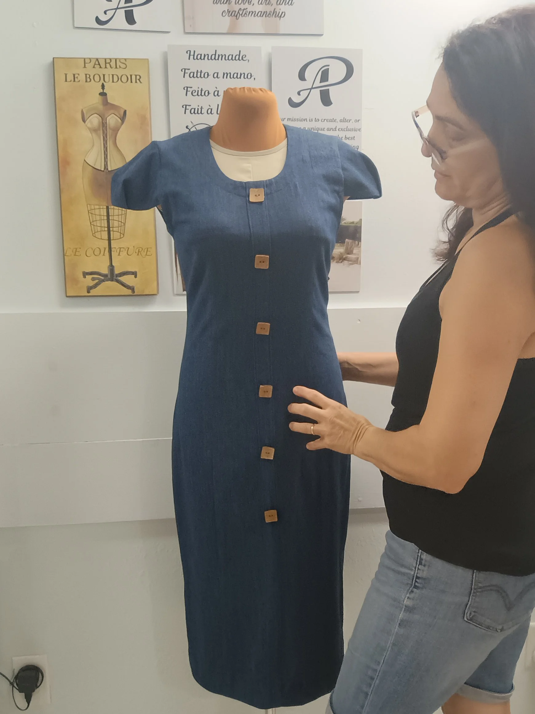 A dress form displaying a denim dress with buttons down the front and puffed sleeves, standing in a room with wall art and signs in the background. A woman is standing next to the dress, touching the lower part of the dress.