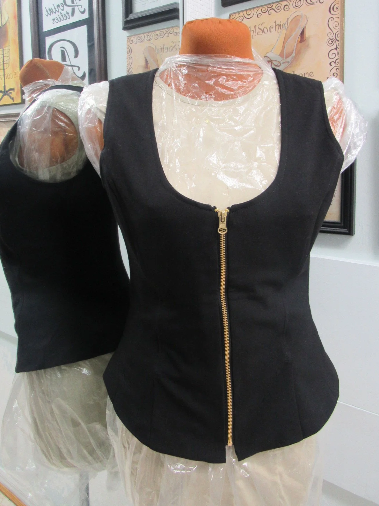 Black sleeveless vest with a front gold zipper, displayed on a mannequin with plastic covering the arms and legs, inside a store with framed posters on the wall in the background.