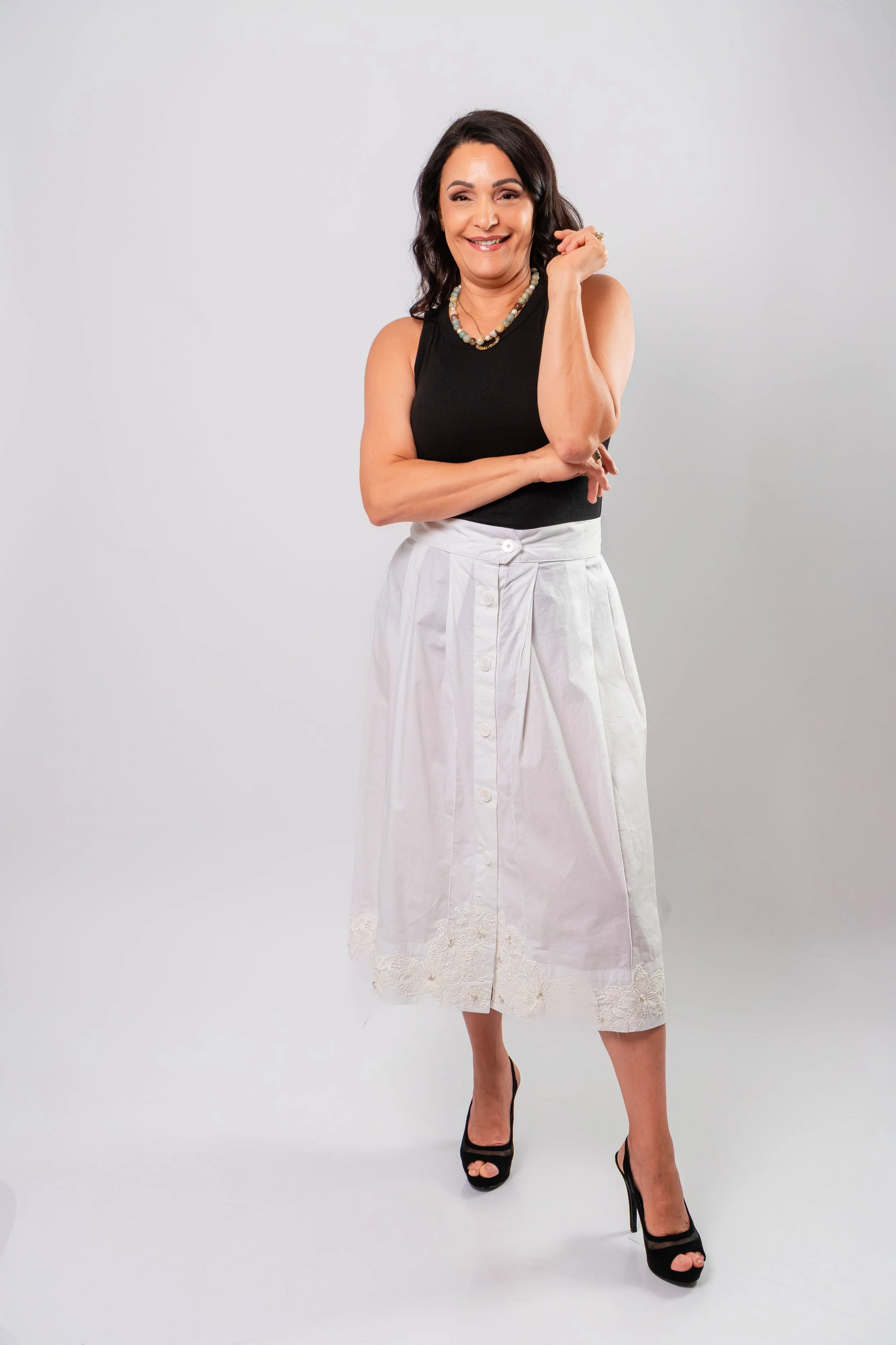 A woman smiling in a studio with a plain background, wearing a black sleeveless top, a white skirt, and black high heels, accessorized with a necklace.