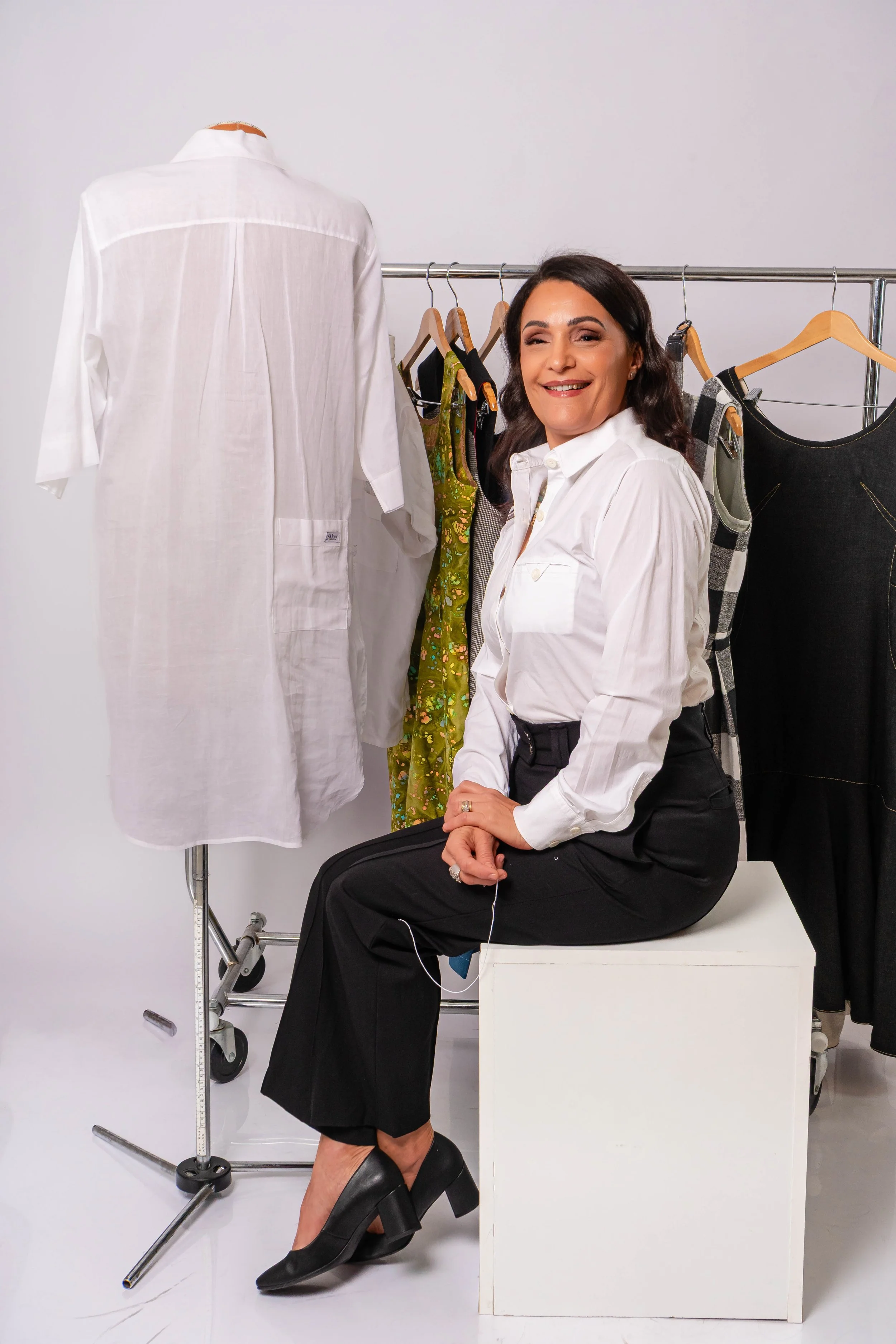 A woman in a white shirt and black pants sitting on a white block in a clothing studio, surrounded by garments on a rack, including a white shirt, a green floral dress, a checked dress, and a black dress.
