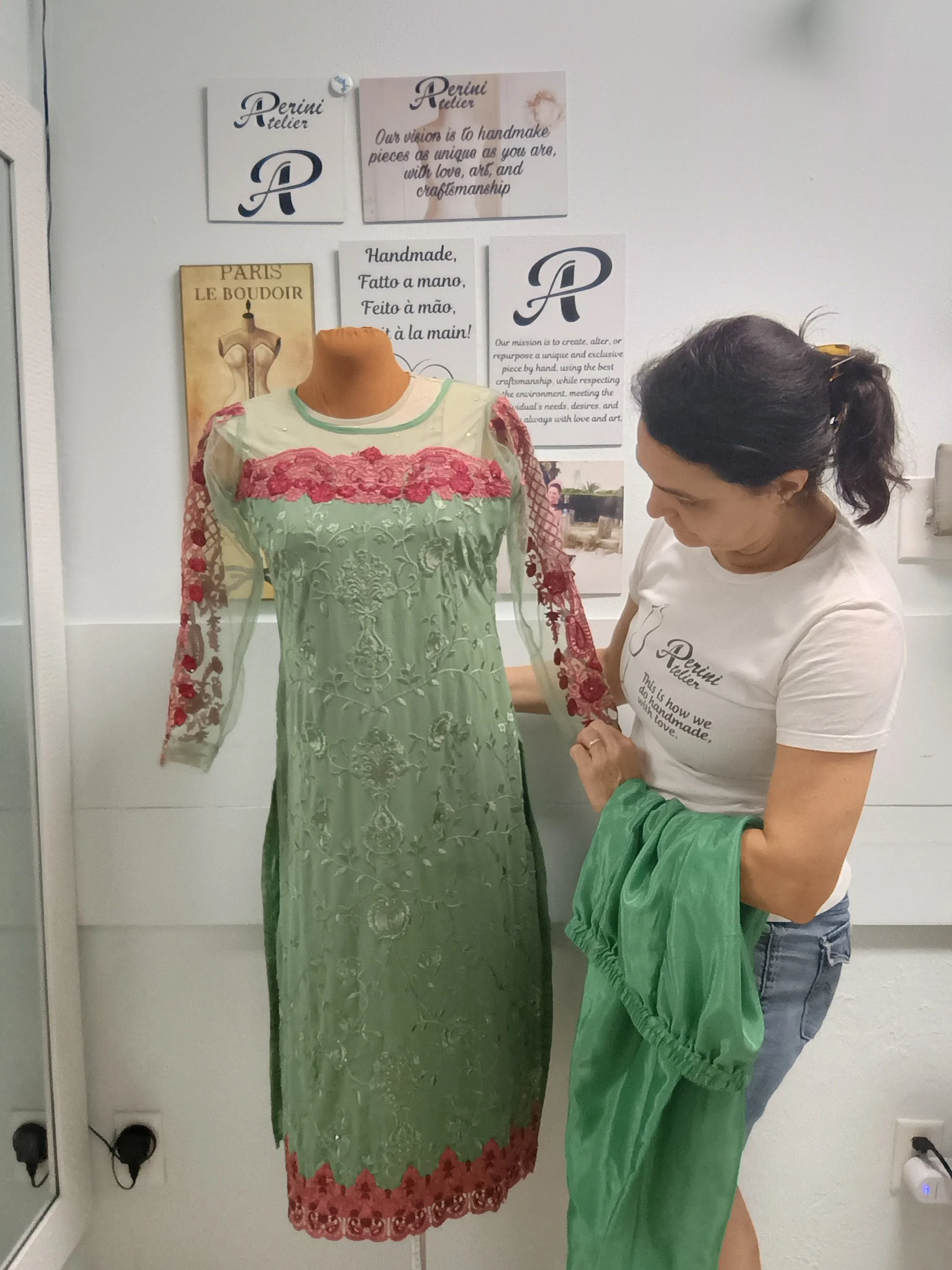 A woman holding a green fabric swatch in front of a mannequin wearing a long, embroidered green dress with red lace accents on the sleeves, neck, and hem. The woman is standing in a room with a white wall decorated with signs about handmade fashion and a logo that reads "Perini Atelier."