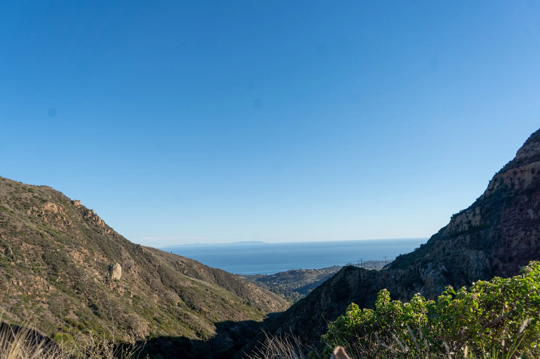 Scenic view of a coastal valley with mountains on either side and the ocean in the distance, under a clear blue sky.