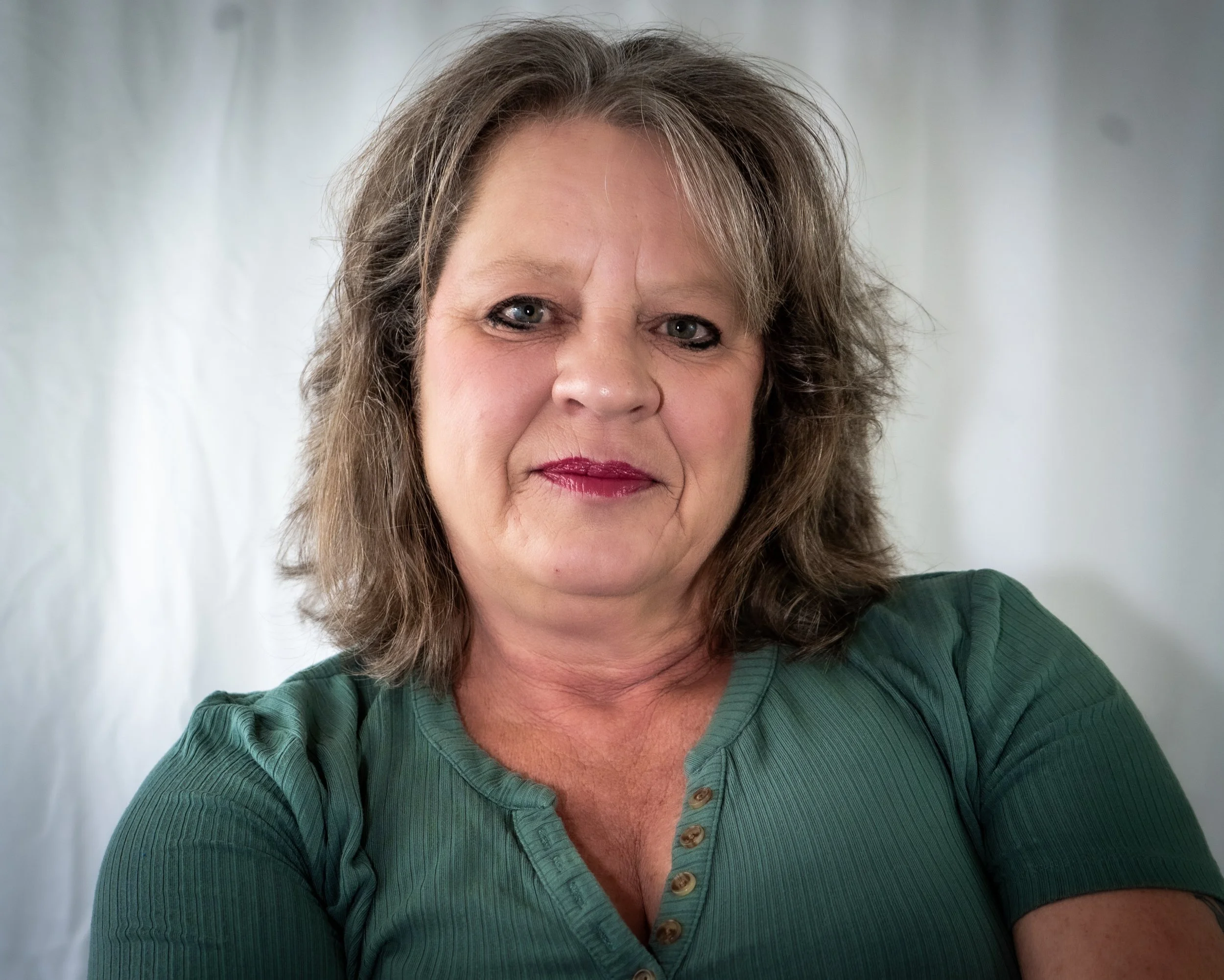 A middle-aged woman with shoulder-length brown hair and blue eyes, wearing a green shirt, posing against a plain white background.
