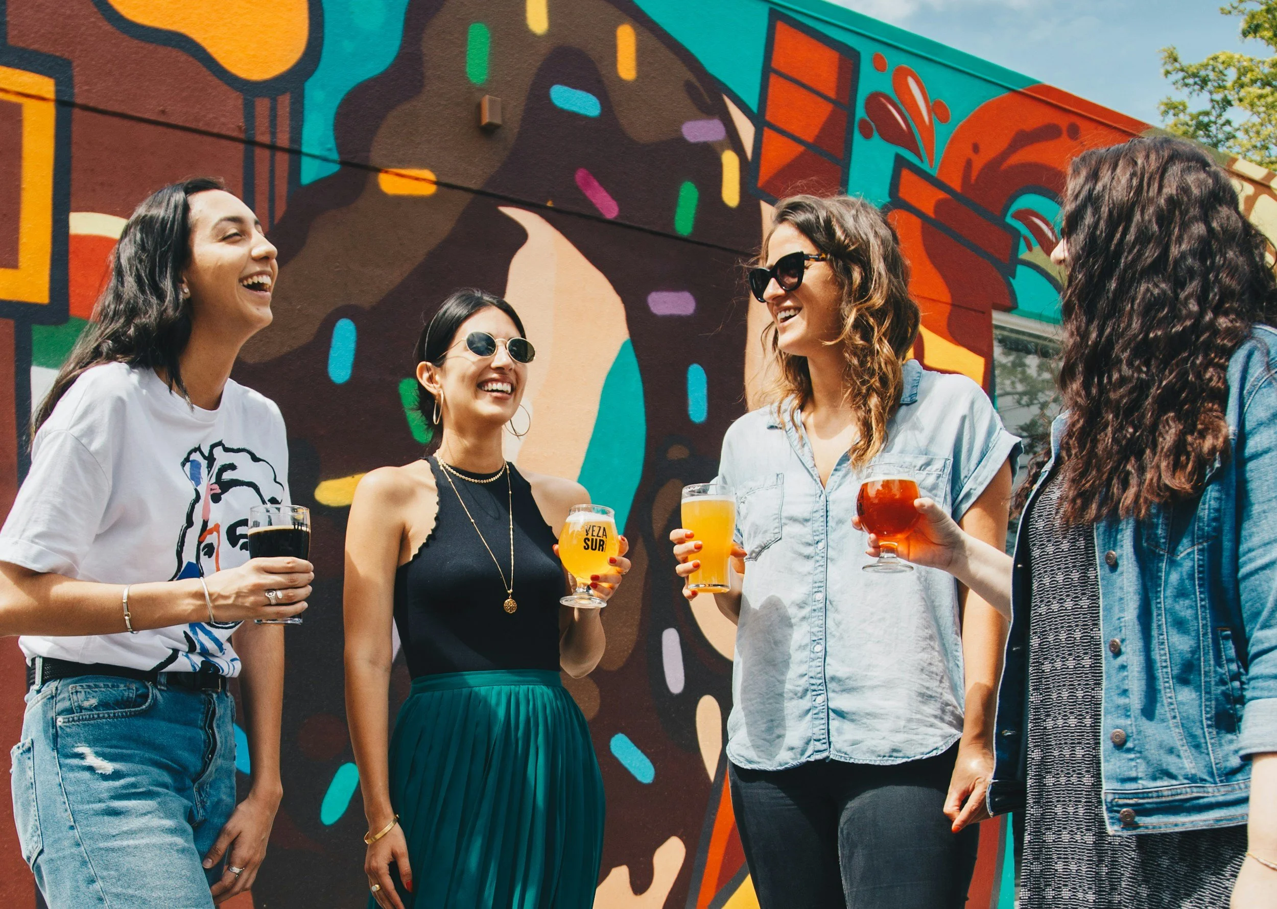 Group of five women standing outdoors, smiling and holding drinks, in front of a colorful mural on a wall.