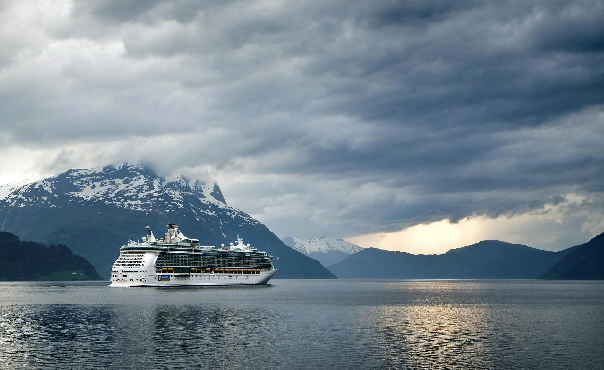 A large cruise ship sailing on calm water with snow-capped mountains and dark, cloudy sky in the background.
