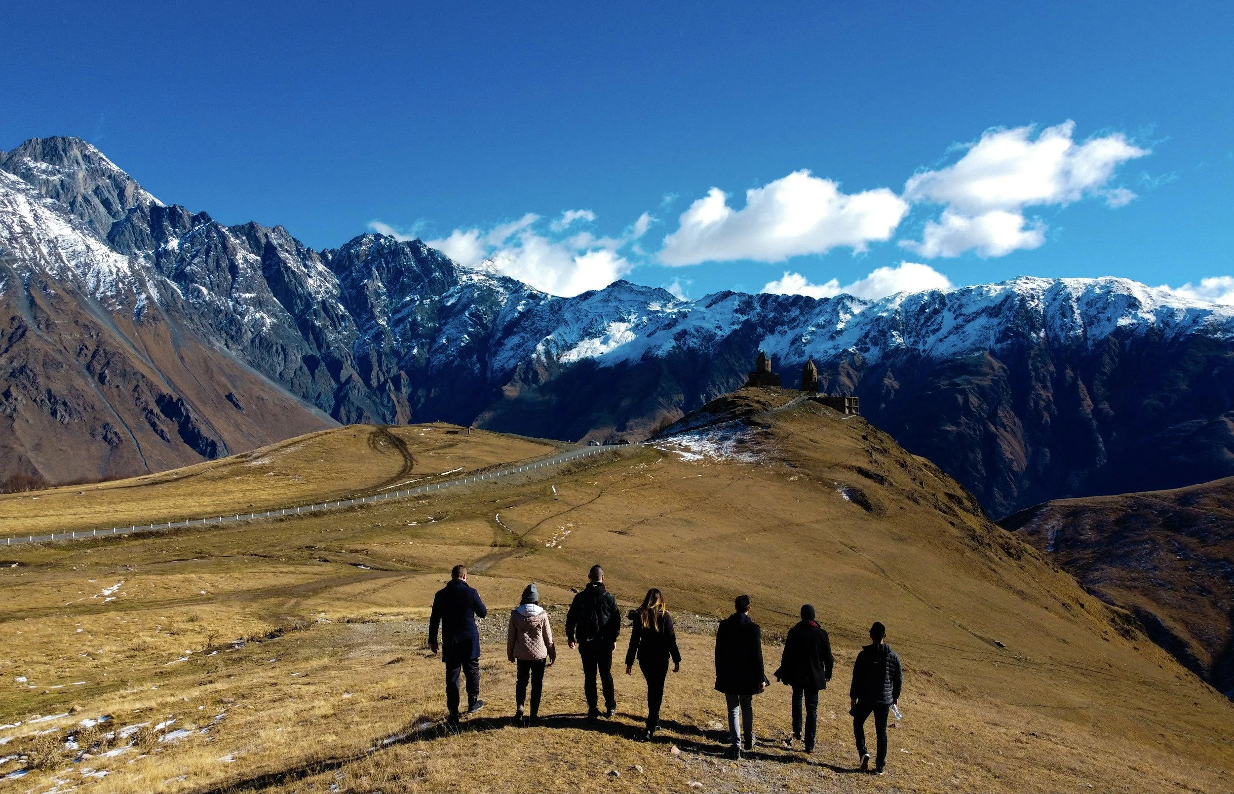 Group of people walking on a trail in a mountainous landscape with snow-capped peaks and blue sky in the background.
