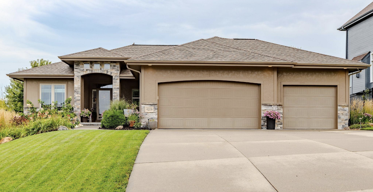 Single-story house with a three-car garage, front yard, green lawn, and landscaping, under a partly cloudy sky.
