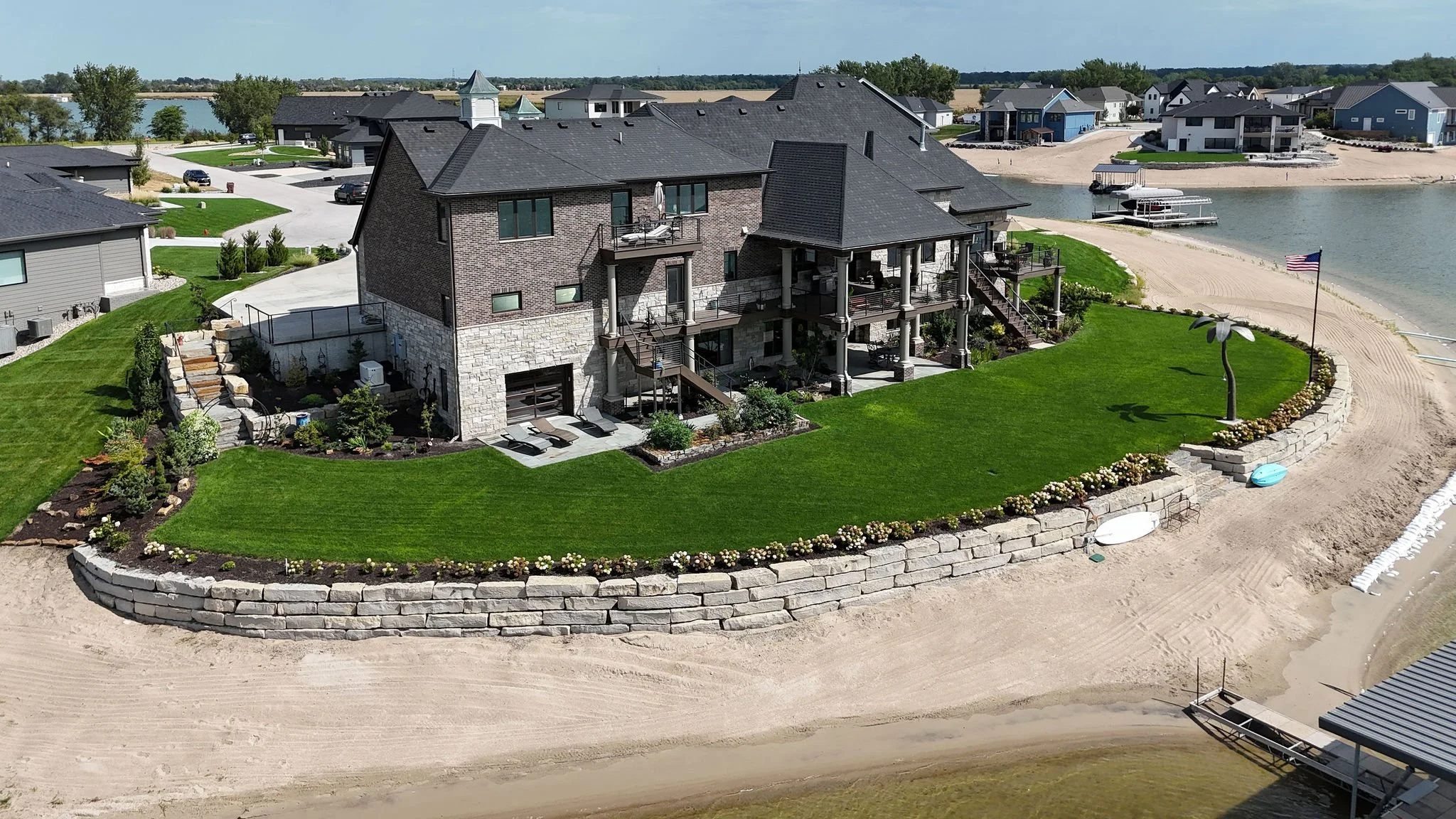 Aerial view of a lakeside house with a landscaped yard, stone retaining wall, and a sandy beach with a pier and boat dock.